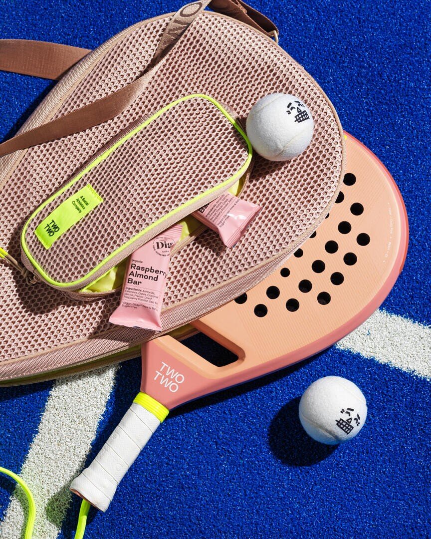 Eine rosa perforierte Paddeltasche, ein rosa Paddel, zwei weiße Bälle, Energieriegel und eine neongrüne Reißverschlusstasche liegen auf einem hellblauen, strukturierten Spielfeld, das mit einer weißen Linie markiert ist.