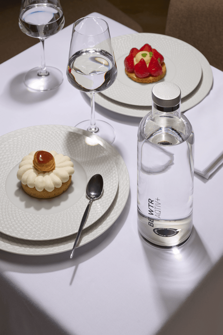 Une table dressée avec deux desserts dans des assiettes blanches, une cuillère, deux verres à eau et une bouteille transparente portant l'inscription "BLK WTR ADITI+" sur une nappe blanche. Un des desserts est recouvert de caramel, l'autre de fraises.