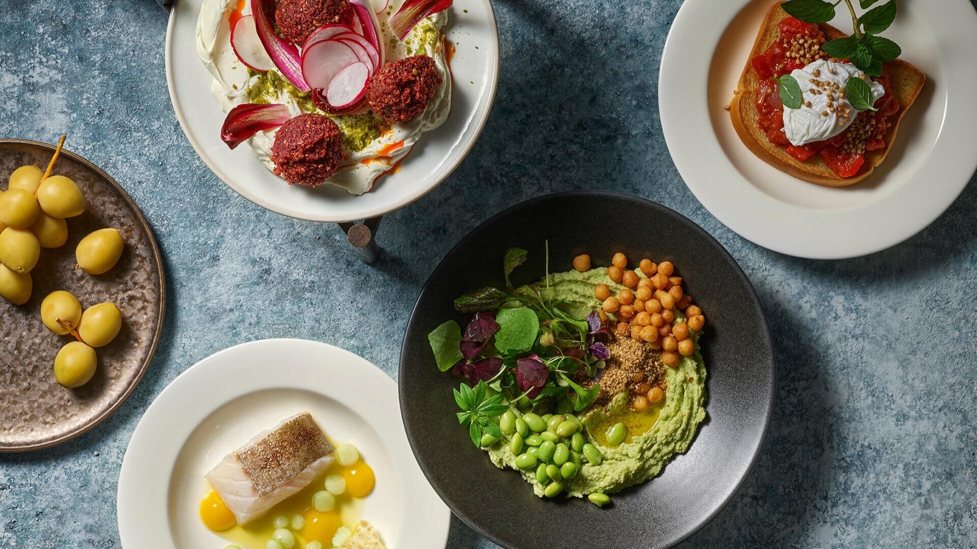 Ansicht von oben auf fünf Teller mit verschiedenen Gerichten, darunter Falafel mit Rettich, ein Dessert mit Sahne, grüner Hummus mit Kichererbsen und Grünzeug, weißer Fisch mit gelber Soße und ein Teller mit gelben Datteln. Alle sind auf einer blau strukturierten Oberfläche angerichtet.