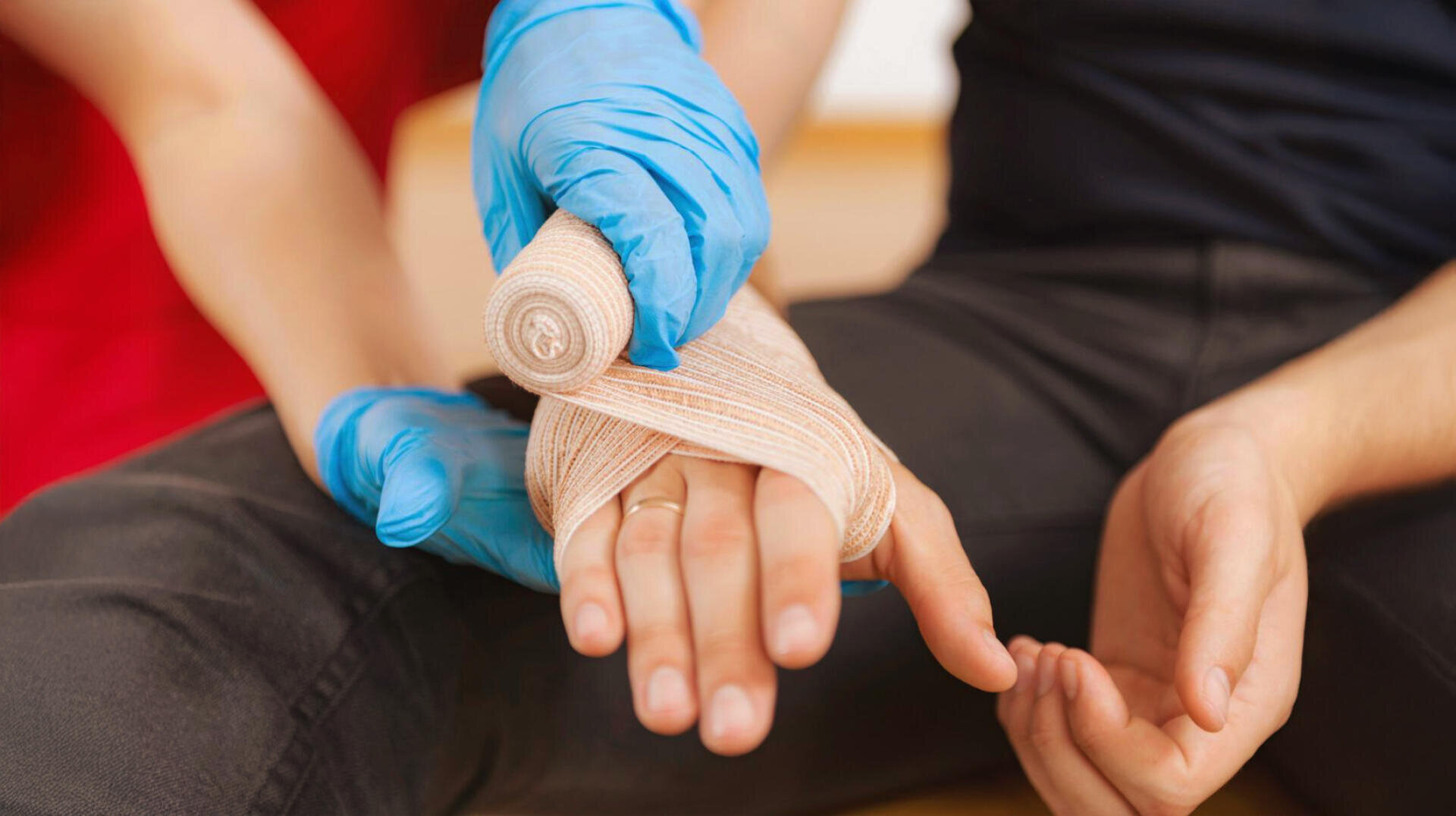 Une personne portant des gants bleus enroule un bandage autour de la main blessée d'une autre personne pendant que celle-ci est assise sur le sol.