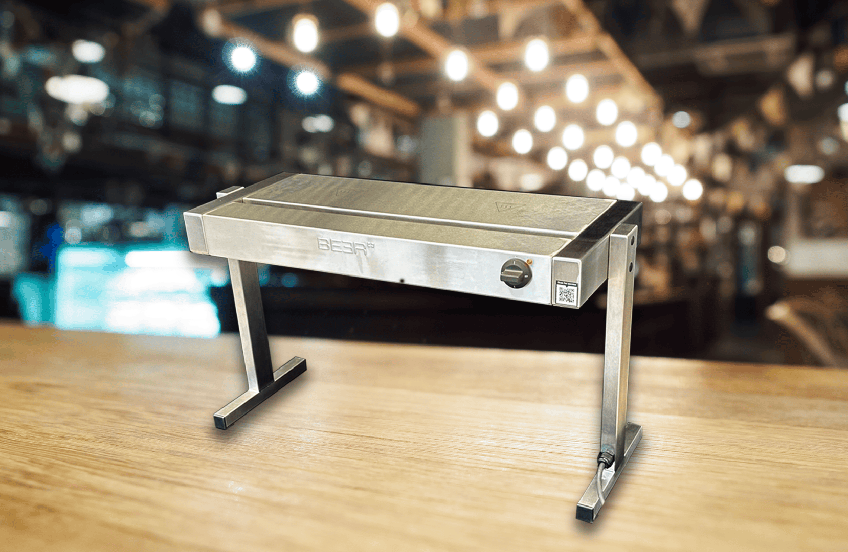 Ein Tischgrill aus rostfreiem Stahl mit Beinen, platziert auf einer hölzernen Oberfläche in einer warm beleuchteten Restaurant- oder Barumgebung. Der Hintergrund ist unscharf und hebt den Grill im Vordergrund hervor.