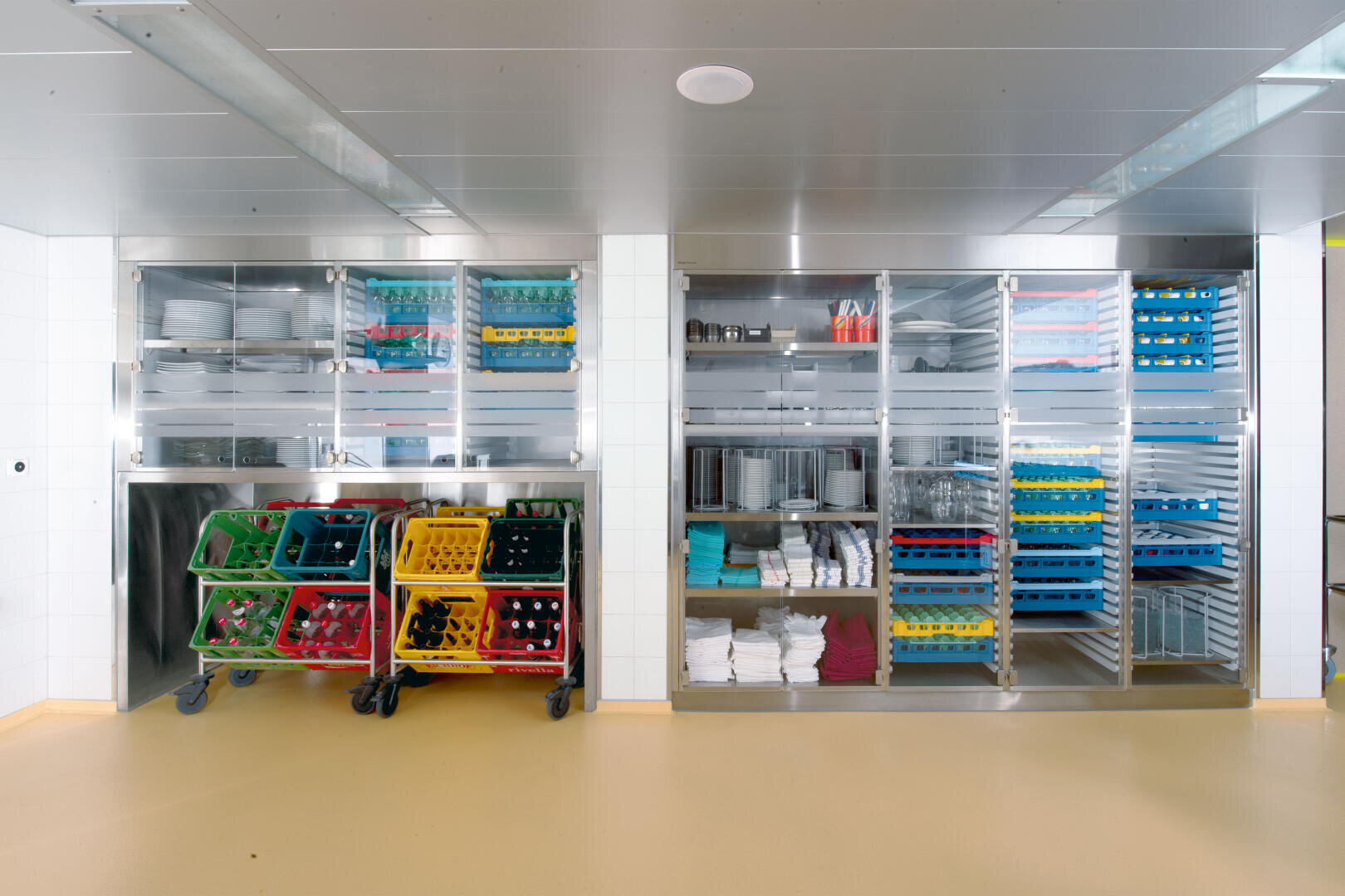 Un espace de rangement propre et organisé dans la cuisine, avec des étagères métalliques contenant des plateaux, des verres, de la vaisselle, des serviettes et des supports de vaisselle colorés sur des chariots à roulettes. La pièce est bien éclairée et le sol est beige.