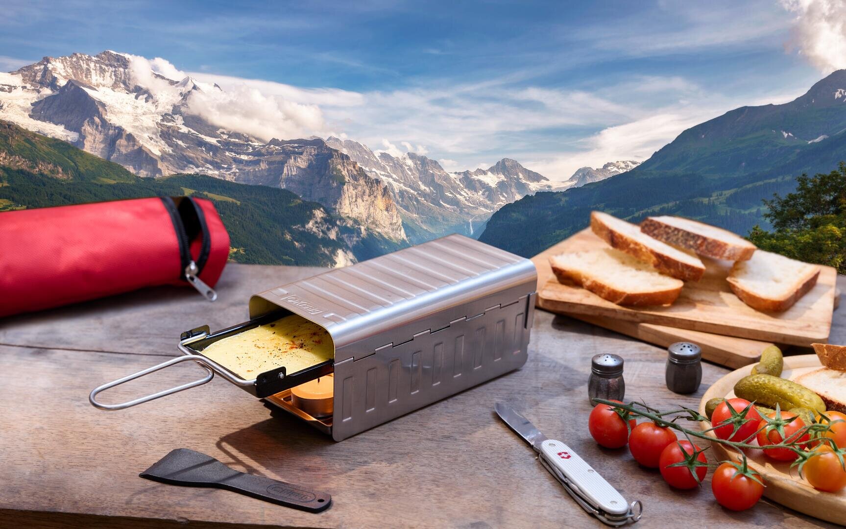 Ein Raclette-Grill aus Metall mit geschmolzenem Käse steht auf einem Holztisch im Freien, umgeben von Brotscheiben, Tomaten, Essiggurken, Salz- und Pfefferstreuern, einem Pfannenwender und einem Schweizer Armeemesser. Im Hintergrund sind schneebedeckte Berge und grüne Täler zu sehen.