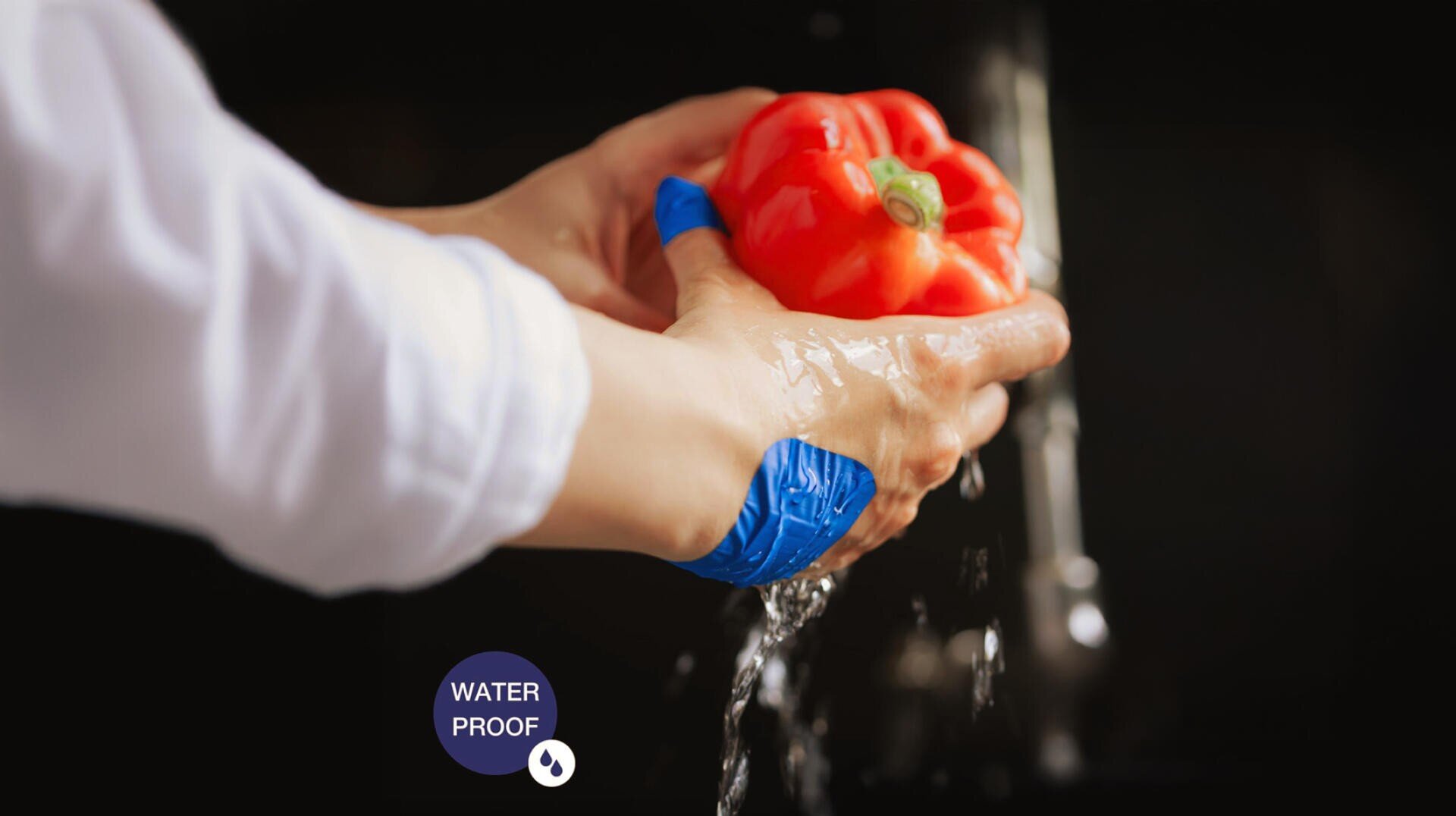 Eine Person wäscht eine rote Paprika unter fließendem Wasser. Ihre Hände sind mit blauen, wasserdichten Verbänden umwickelt, in der unteren linken Ecke ist ein „WASSERDICHT“-Schild zu sehen.