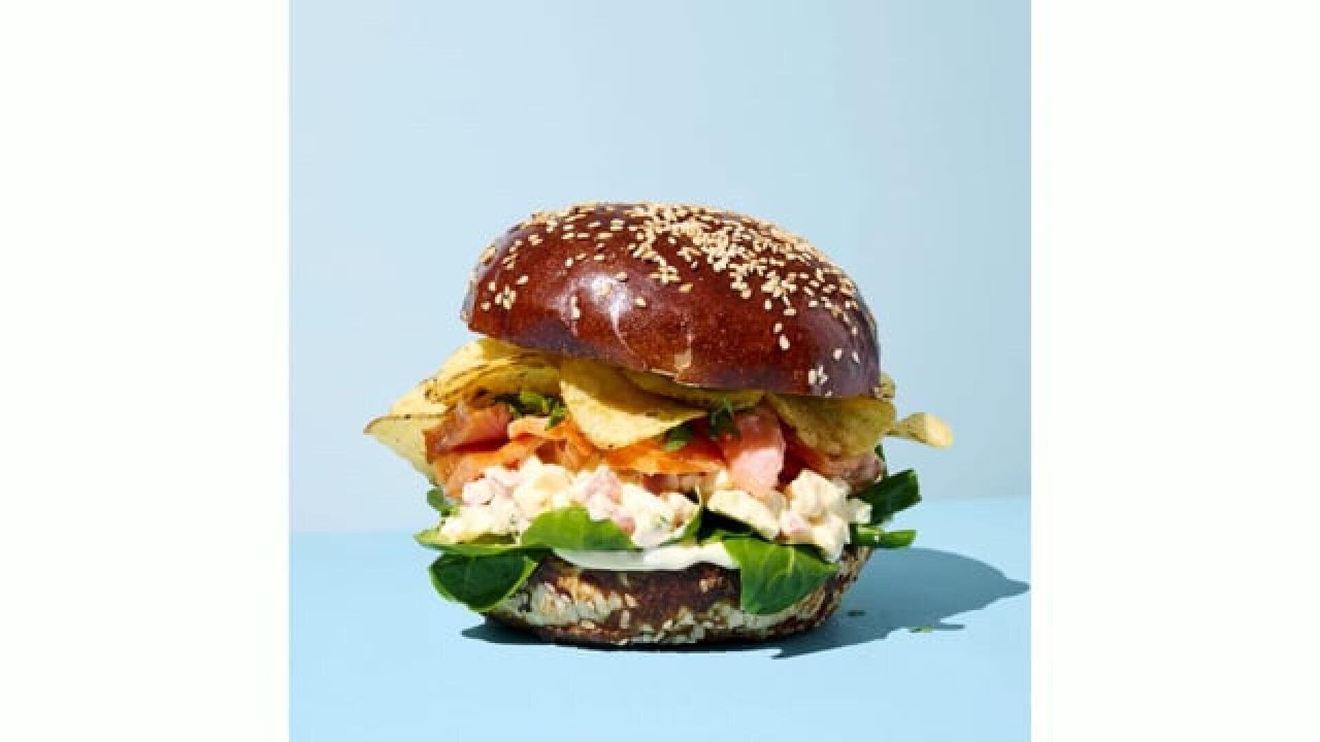 Ein Burger mit einem Laugen-Sesambrötchen, Blattgemüse, knackigem Salat, Tomatenscheiben, Kartoffelchips und Lachsstückchen vor einem hellblauen Hintergrund.