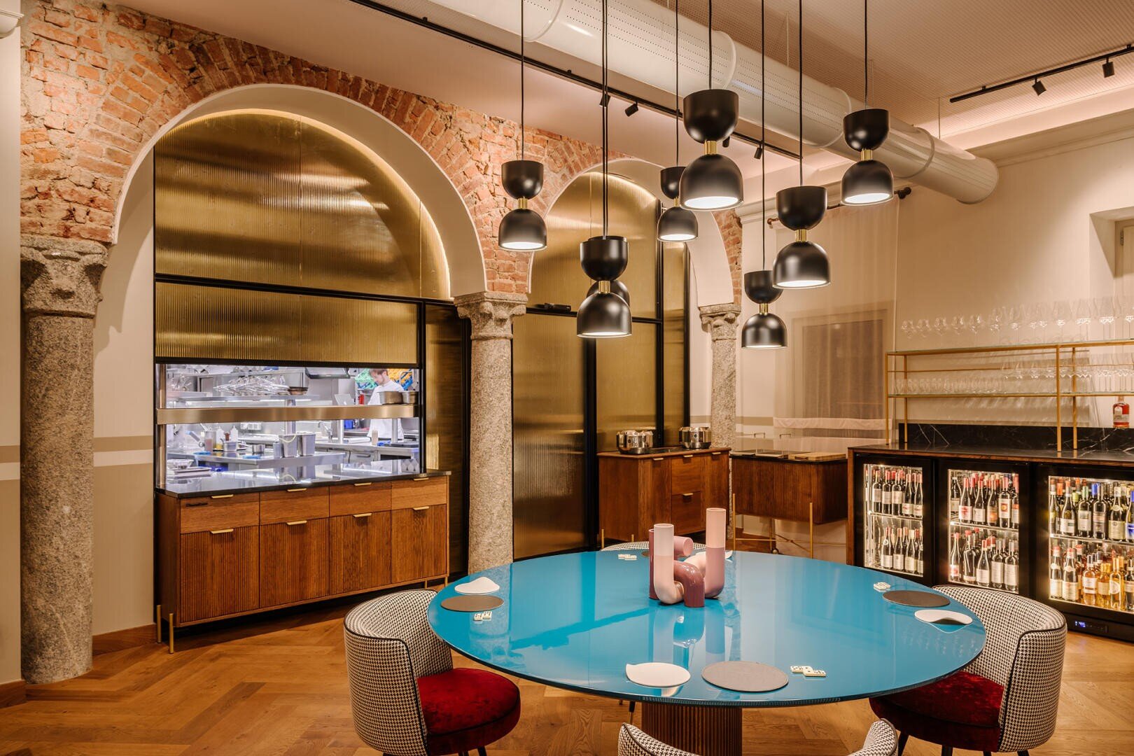 Une salle à manger moderne et élégante avec des arches en briques apparentes, une table ronde turquoise, des lampes suspendues contemporaines, des chaises rembourrées, des verres sur des étagères, un réfrigérateur à vin et une vue sur une cuisine élégante.