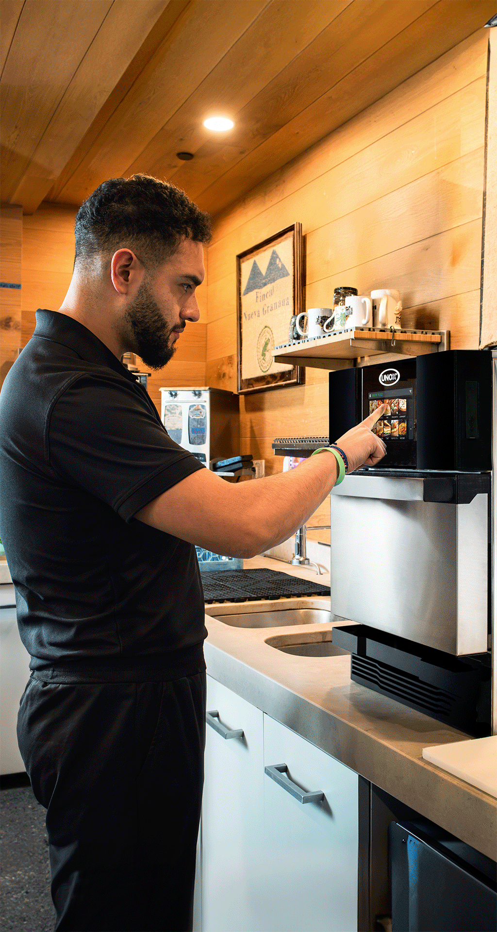 Un homme en chemise noire utilise une machine à café à écran tactile dans une cuisine moderne avec des murs lambrissés, des armoires blanches et divers articles de cuisine sur le comptoir et les étagères.