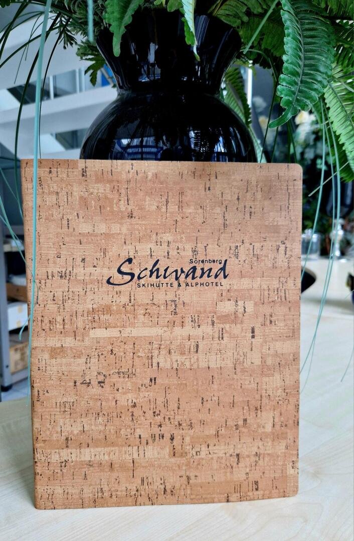 Eine mit Kork überzogene Speisekarte mit der Aufschrift Schivana Skihütte Alphotell steht aufrecht auf einem Tisch mit einer schwarzen Vase und Grünpflanzen im Hintergrund.