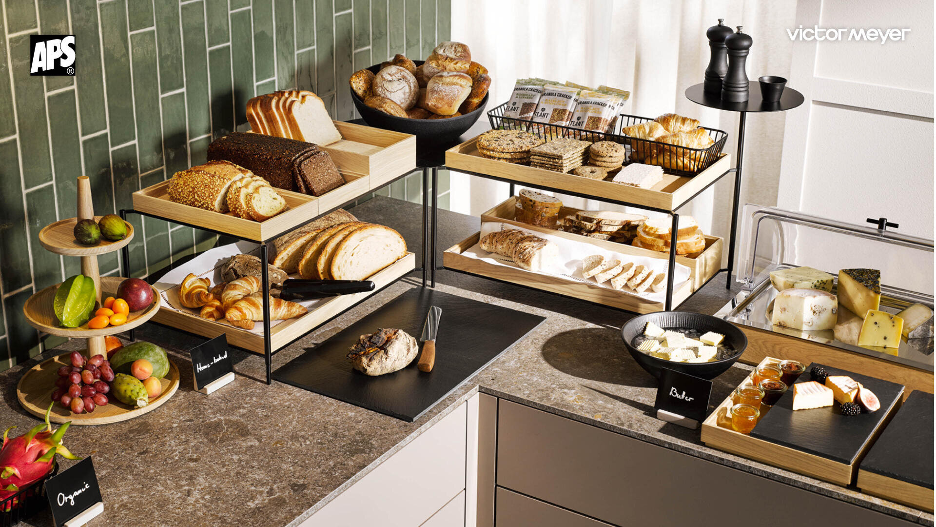 Un buffet avec différentes sortes de pains, de petits pains, de pâtisseries, de fruits, de fromages et d'épices, des ustensiles de service et des panneaux étiquetés se trouve devant un mur carrelé vert dans une cuisine moderne.