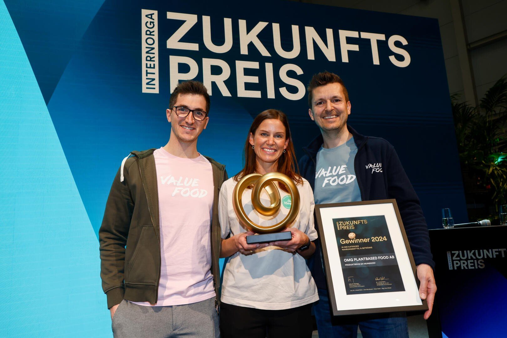 Drei lächelnde Menschen stehen bei der Verleihung des INTERNORGA Zukunftspreises gemeinsam auf der Bühne. Die Frau in der Mitte hält eine goldene Spiral-Trophäe, der Mann rechts hält eine gerahmte Urkunde.