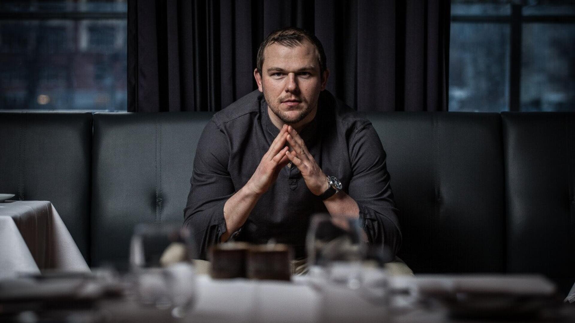 Un homme est assis sur une banquette sombre dans un restaurant, penché en avant, les mains serrées l'une contre l'autre, regardant directement la caméra. La table devant lui est garnie de verres et d'assiettes, et l'arrière-plan comporte de grandes fenêtres.
