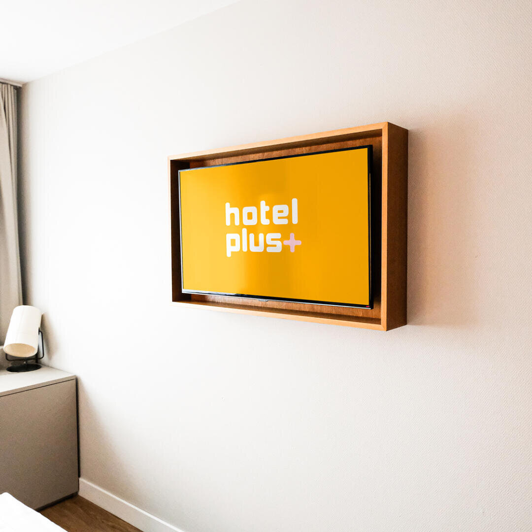 Ein auf einem Holzrahmen montierter Flachbildfernseher zeigt einen leuchtend gelben Bildschirm mit der Aufschrift hotel plus+ in weißer Schrift. Der Fernseher steht an einer hellen Wand in einem modernen Hotelzimmer.