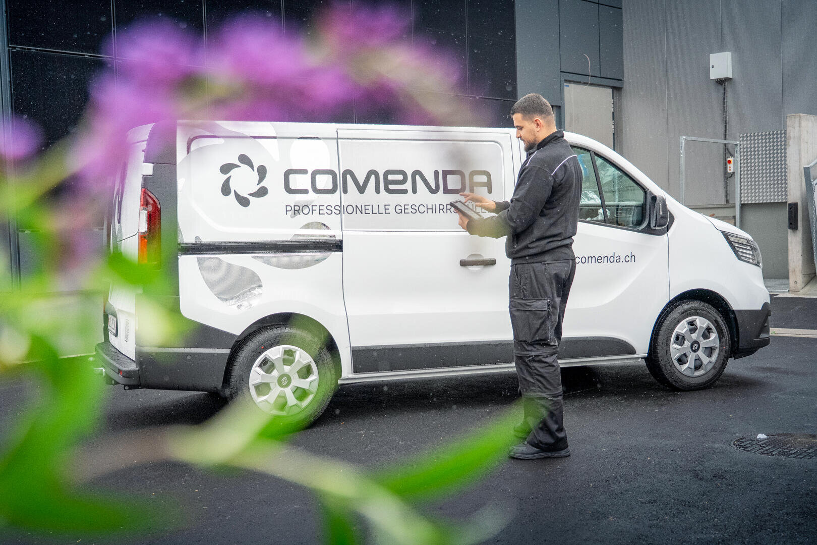 Ein Mann in schwarzer Arbeitskleidung steht neben einem weißen Lieferwagen der Firma Comenda und schreibt auf einem Klemmbrett. Die Szene spielt sich im Freien ab, mit einer verschwommenen lila Blume im Vordergrund.