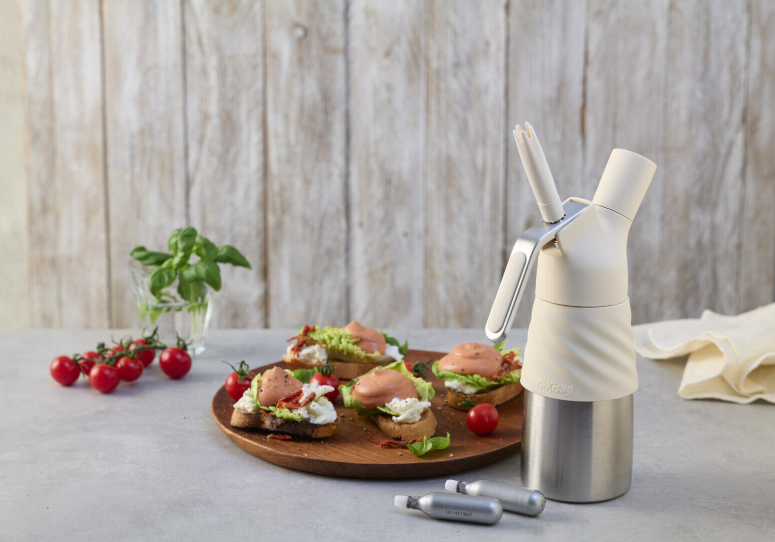 Un siphon à crème est posé sur une table, à côté de deux cartouches de gaz, et un plateau en bois garni de pain grillé, d'avocat, de crème et de saumon fumé. Des tomates cerises et du basilic sont à l'arrière-plan.