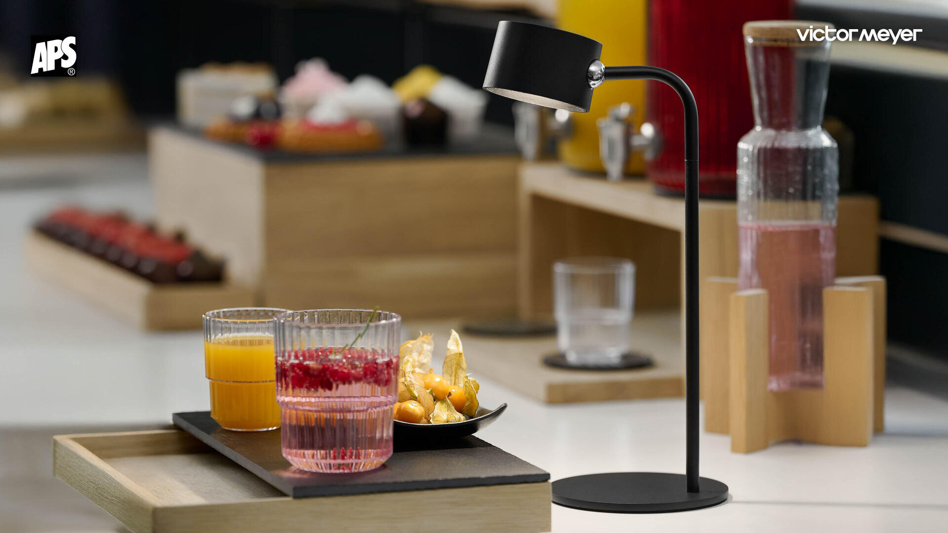 Une lampe de table moderne éclaire un plateau avec deux verres de jus de fruits et une petite assiette de snacks sur une table blanche. D'autres aliments et boissons sont flous en arrière-plan.
