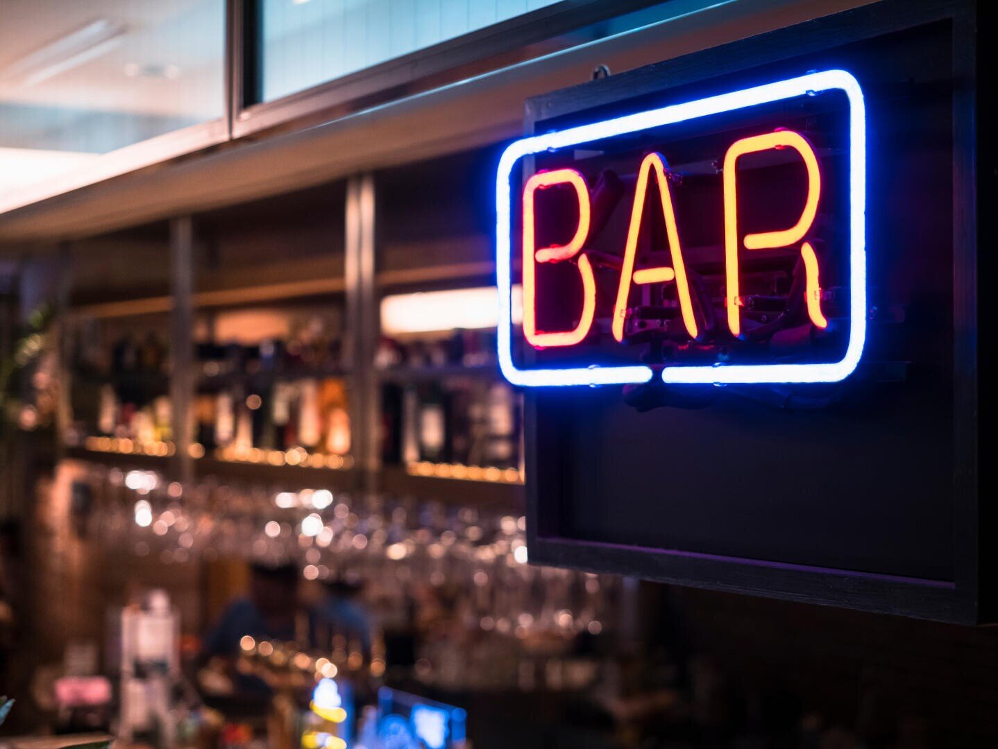 Ein hell leuchtendes Neonschild mit der Aufschrift BAR hängt in einer schwach beleuchteten Bar, in der im Hintergrund Flaschen und Gläser zu sehen sind.