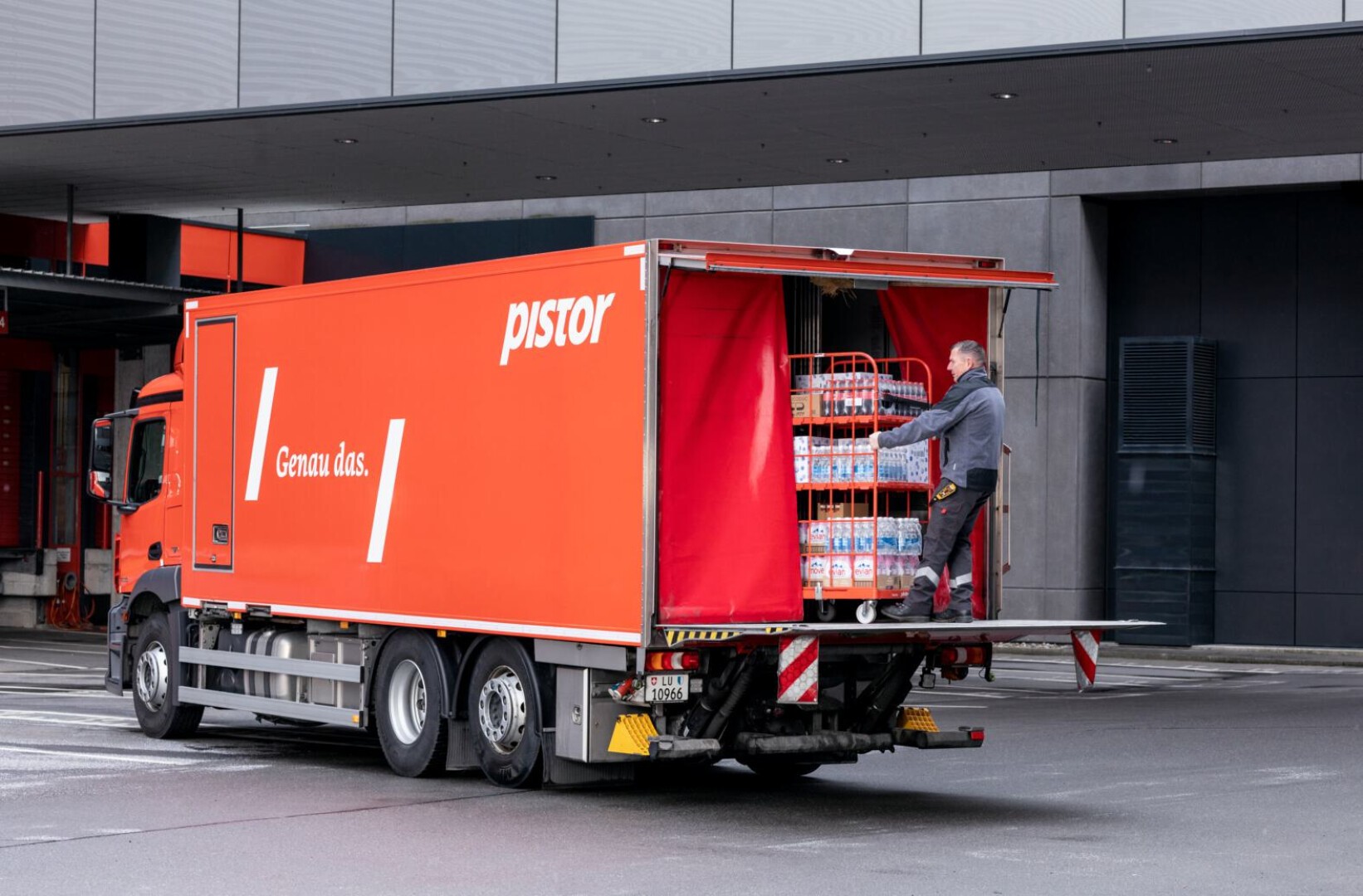 Ein Lieferwagen mit Pistor-Aufdruck und einem Slogan steht vor einem Gebäude. Ein Mann steht auf der Hebebühne und lädt einen Wagen voller Getränkeflaschen aus.