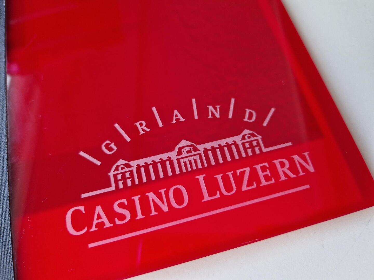 Eine rote Acryltafel, auf der der Schriftzug Grand Casino Luzern und die Abbildung eines Gebäudes mit drei spitzen Dächern eingraviert sind.