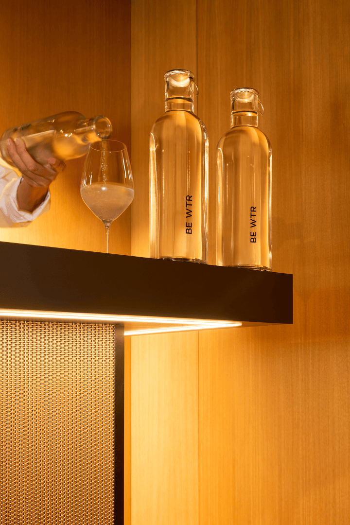 Une personne verse une boisson d'une bouteille transparente dans un verre à vin sur un comptoir de bar moderne. Deux hautes bouteilles transparentes portant l'inscription BE VITR sont placées à côté du verre, le tout sur un fond de bois chaleureux.
