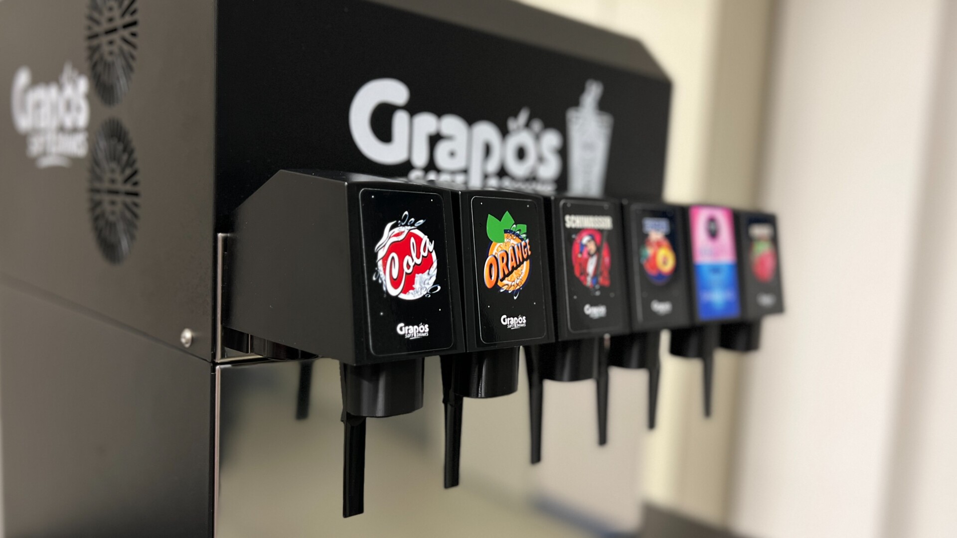 Nahaufnahme eines Grapos-Getränkeautomaten mit fünf Getränkeoptionen, jeweils mit buntem Logo und schwarzen Hebeln zur Getränkeausgabe. Der Hintergrund ist unscharf.