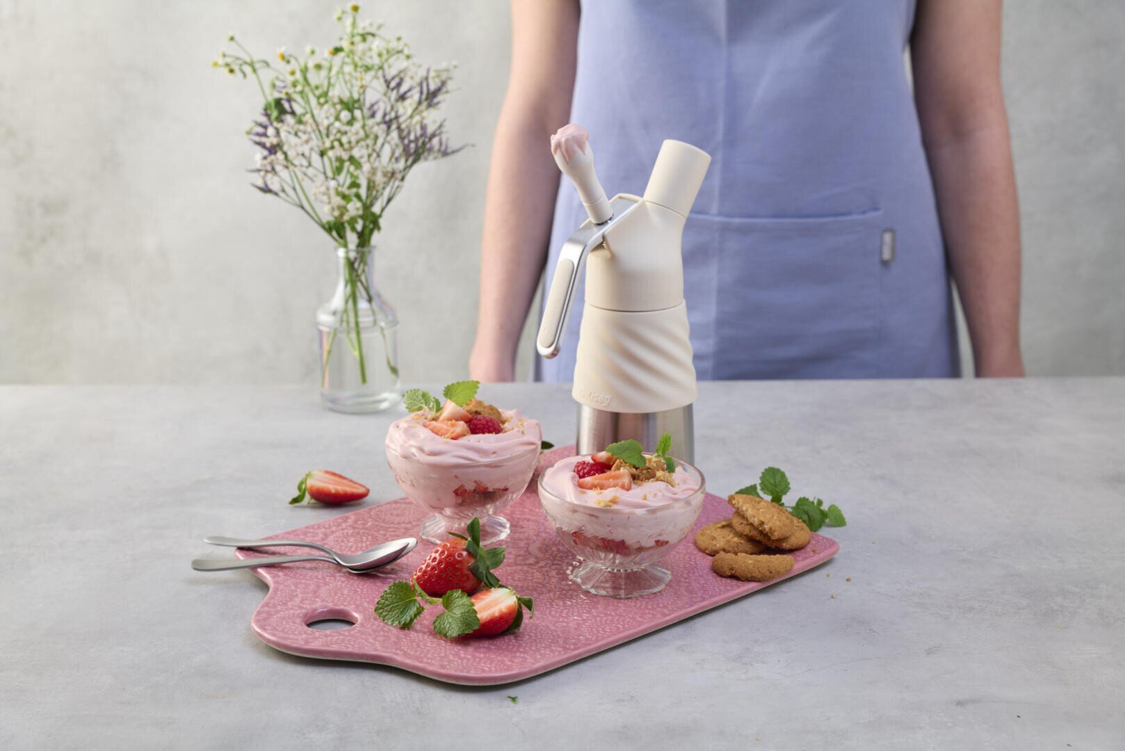Eine Person in einer hellblauen Schürze steht hinter einem Tisch mit zwei Schüsseln Erdbeermousse, garniert mit Minze, Erdbeeren und Keksen. Ein Sahnespender, Löffel und eine kleine Vase mit Blumen stehen ebenfalls auf dem Tisch.