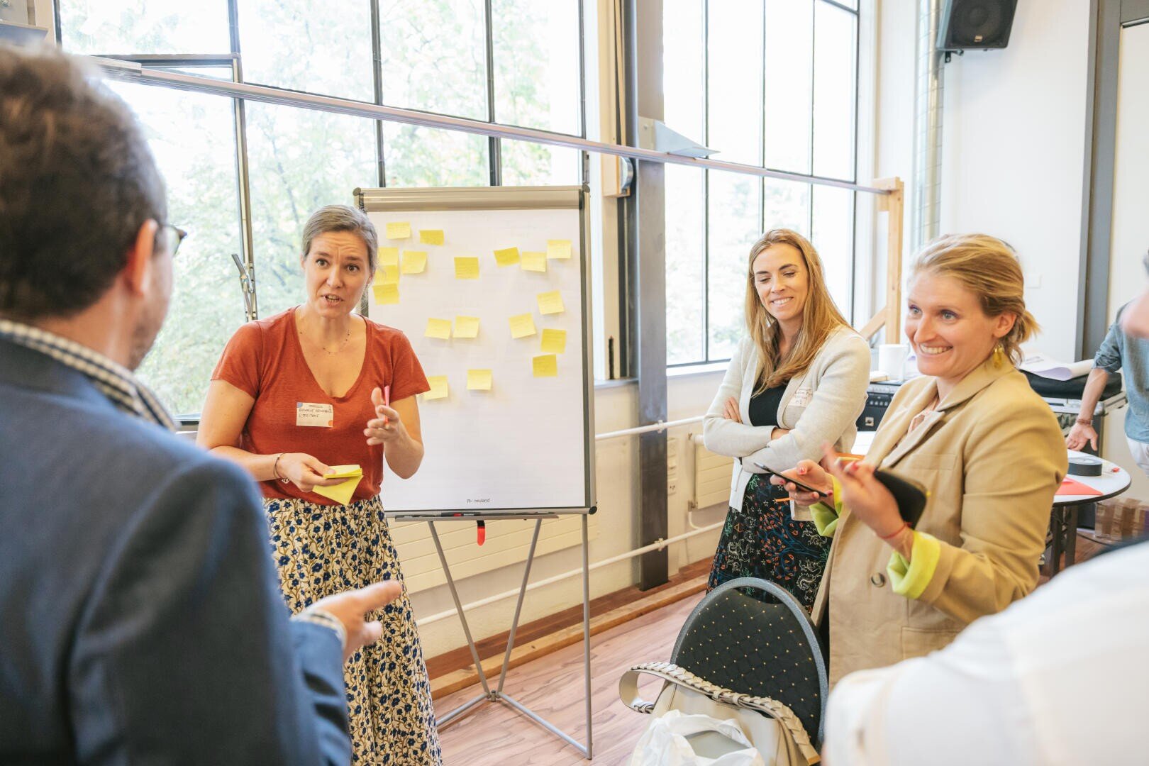 Vier Personen unterhalten sich in einem hellen Raum um ein Flipchart mit Haftnotizen. Drei Frauen halten Notizblöcke in der Hand und hören einem Mann beim Sprechen zu. Im Hintergrund sind große Fenster und Bäume zu sehen.