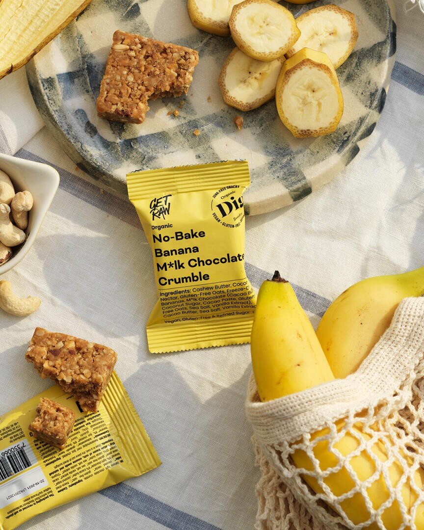 Eine flache Lage Bananen, Bananenscheiben auf einem Teller, Cashewnüsse und ein gelber Get Raw No-Bake Banana M*lk Chocolate Crumble Snackriegel, dessen Verpackung auf einem gestreiften Tuch zu sehen ist.