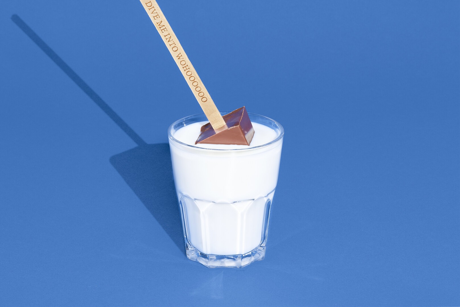 Ein Glas Milch auf einer blauen Oberfläche mit einem Holzstab, der ein pyramidenförmiges Stück Schokolade hält, das teilweise in die Milch eingetaucht ist.