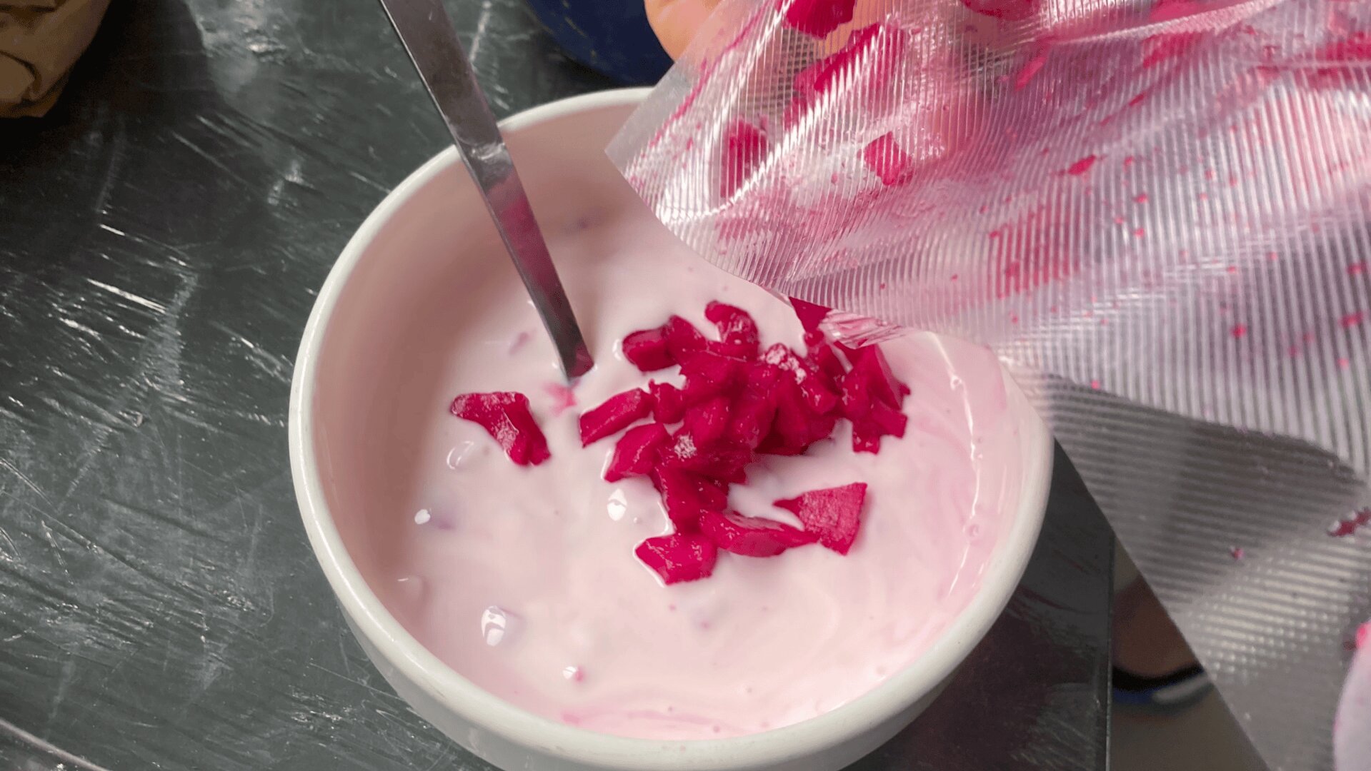 Eine Hand schüttet gehacktes, hellrosa eingelegtes Gemüse aus einem Plastikbeutel in eine Schüssel mit hellrosa Joghurt, mit einem Metalllöffel in der Schüssel, auf einer metallischen Oberfläche.