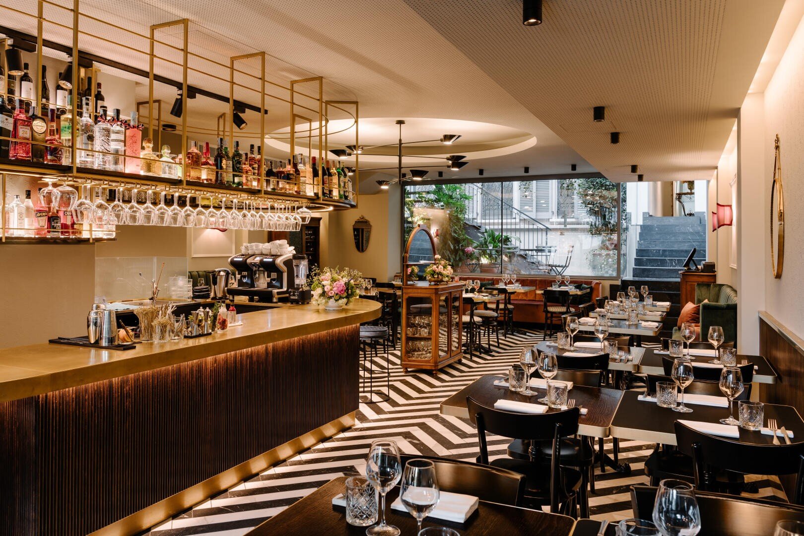 Intérieur d'un restaurant moderne avec un bar élégant, de la verrerie et des bouteilles sur des étagères. Les tables noires sont garnies de verres et d'assiettes ; le sol en zigzag mène à de grandes fenêtres à l'arrière, qui laissent entrer la lumière naturelle.