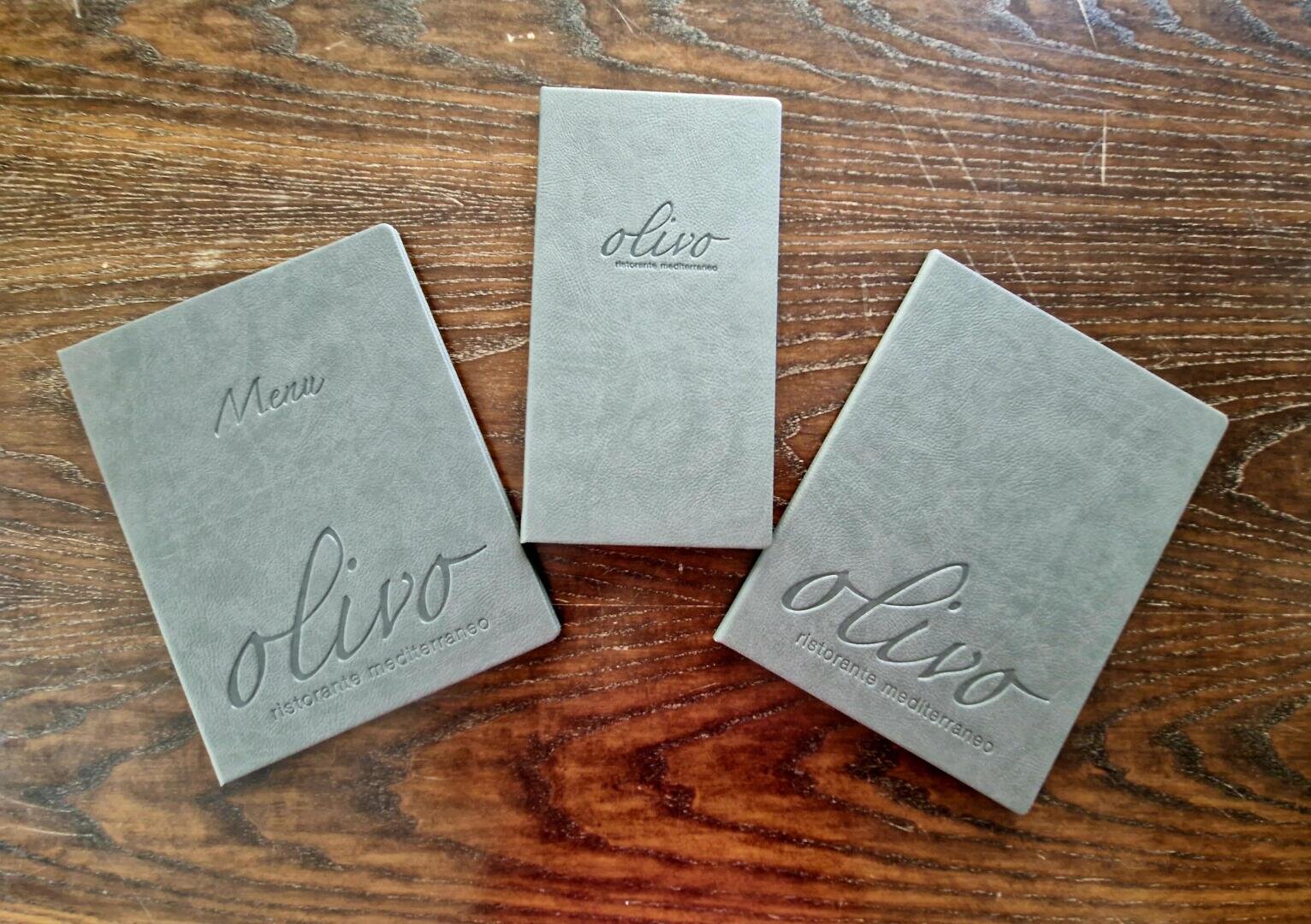 Auf einem Holztisch liegen drei graue Speisekartenhefte mit der Aufschrift olivo ristorante mediterraneo. Auf dem linken Heft steht ebenfalls Menu.