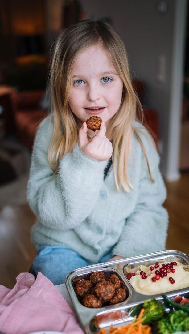 Ein junges Mädchen mit langen blonden Haaren und einem hellblauen Pullover sitzt an einem Tisch und hält eine vegane Frikadelle an ihren Mund. Vor ihr steht ein Essenstablett mit Bits for Kids, Kartoffelpüree, Preiselbeeren und Gemüse.