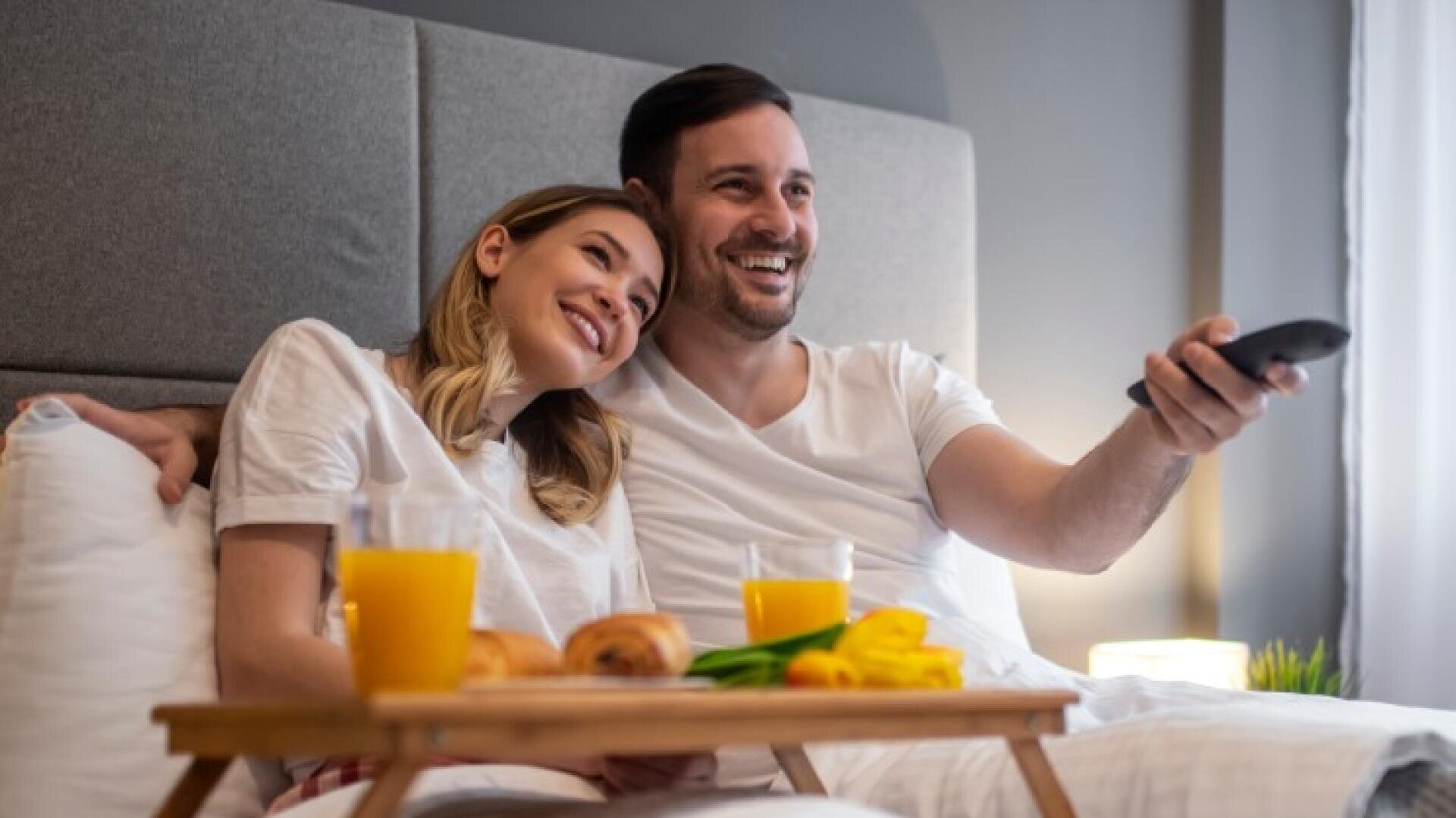 Ein Paar sitzt auf einem Bett, lächelt und sieht fern. Vor ihnen steht das Frühstück auf einem Tablett mit Orangensaft, Croissants und gelben Blumen. Beide tragen weiße Hemden und sehen entspannt und glücklich aus.