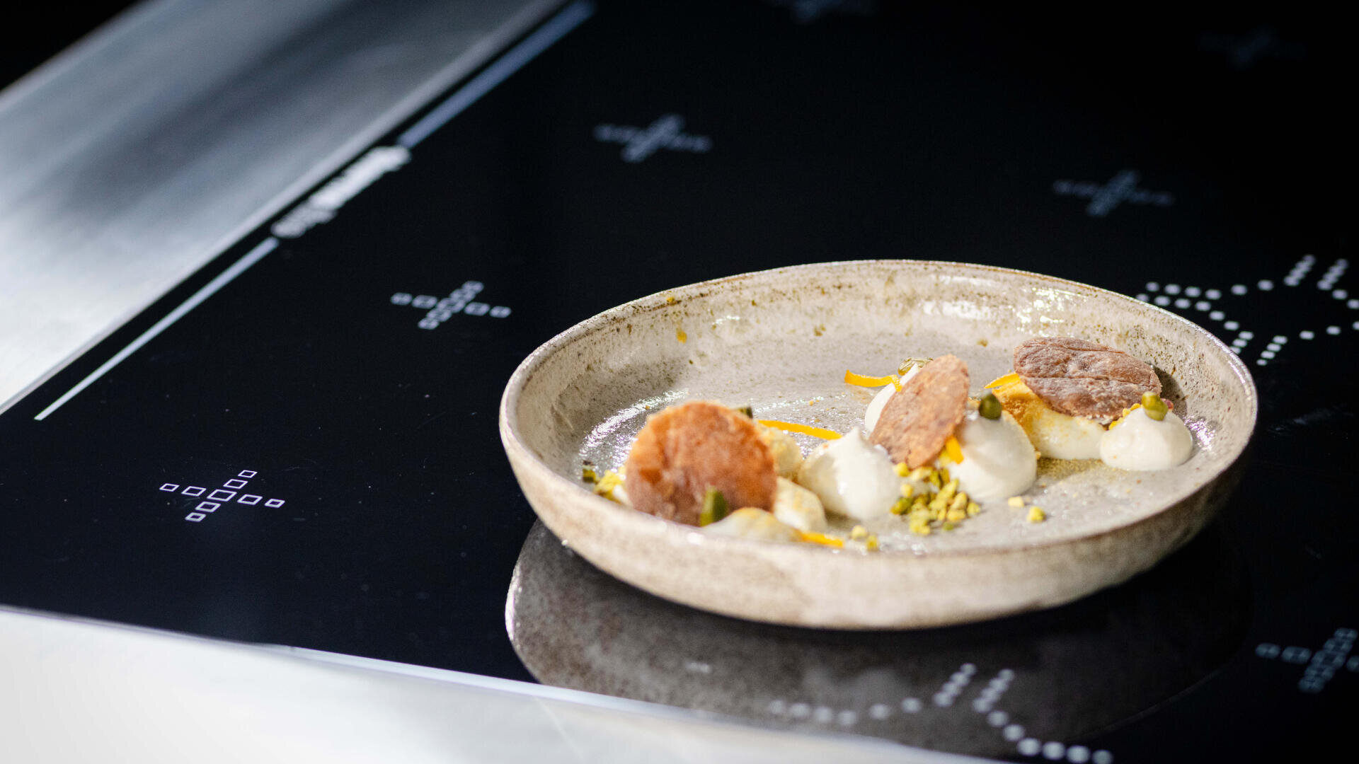 Ein Keramikteller mit einem Gourmet-Dessert zeigt kleine Löffel Sahne, zwei runde Gebäckstücke und vereinzelte gelbe Garnituren, alles auf einem eleganten schwarzen Induktionskochfeld.