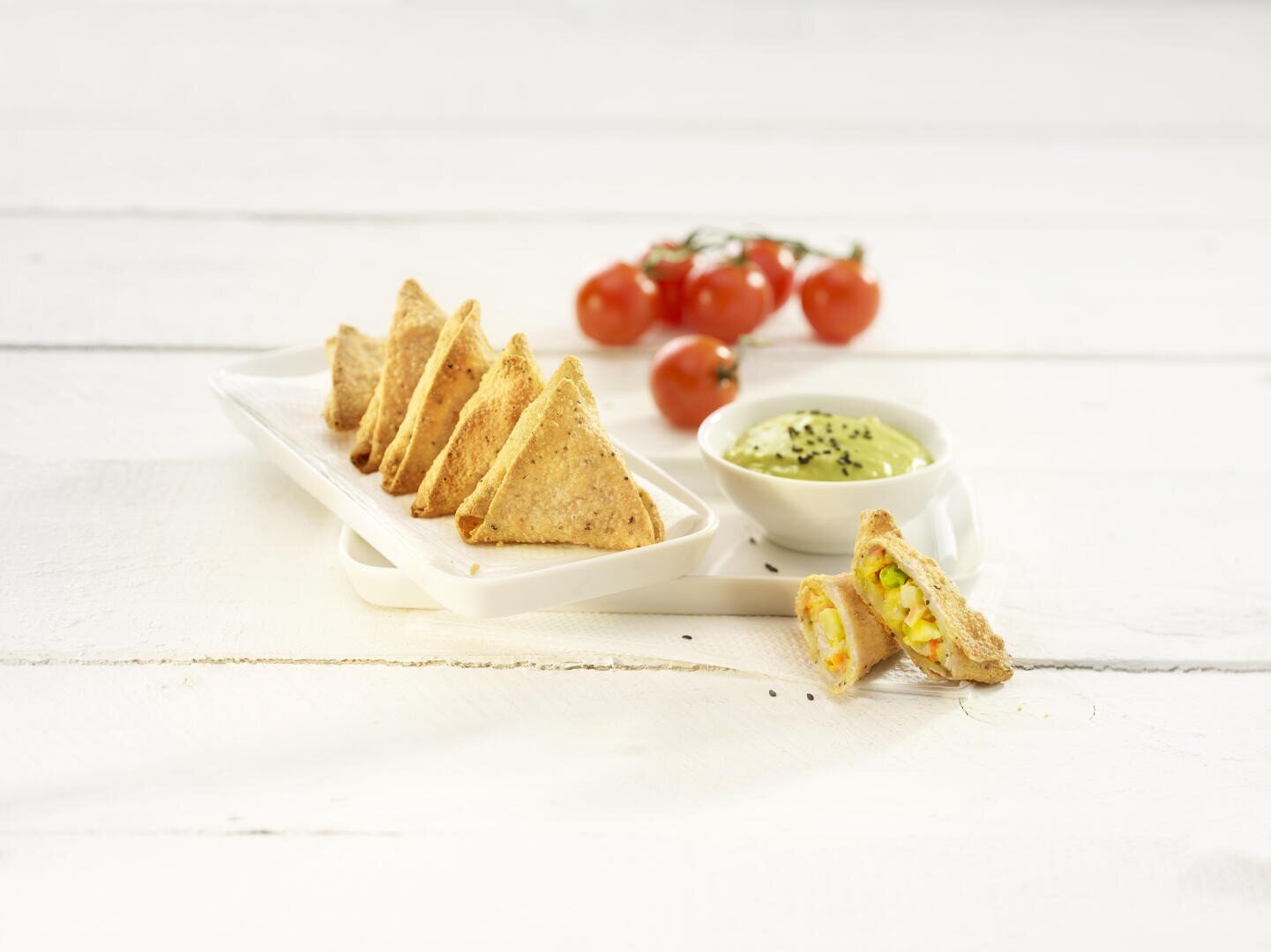 Eine Reihe dreieckiger, goldener Snacks auf einem weißen Tablett, von denen einer geöffnet ist, um eine Gemüsegarnitur zu zeigen. Eine Schüssel mit grüner Soße mit schwarzen Samen und eine Traube Kirschtomaten befinden sich im Hintergrund.