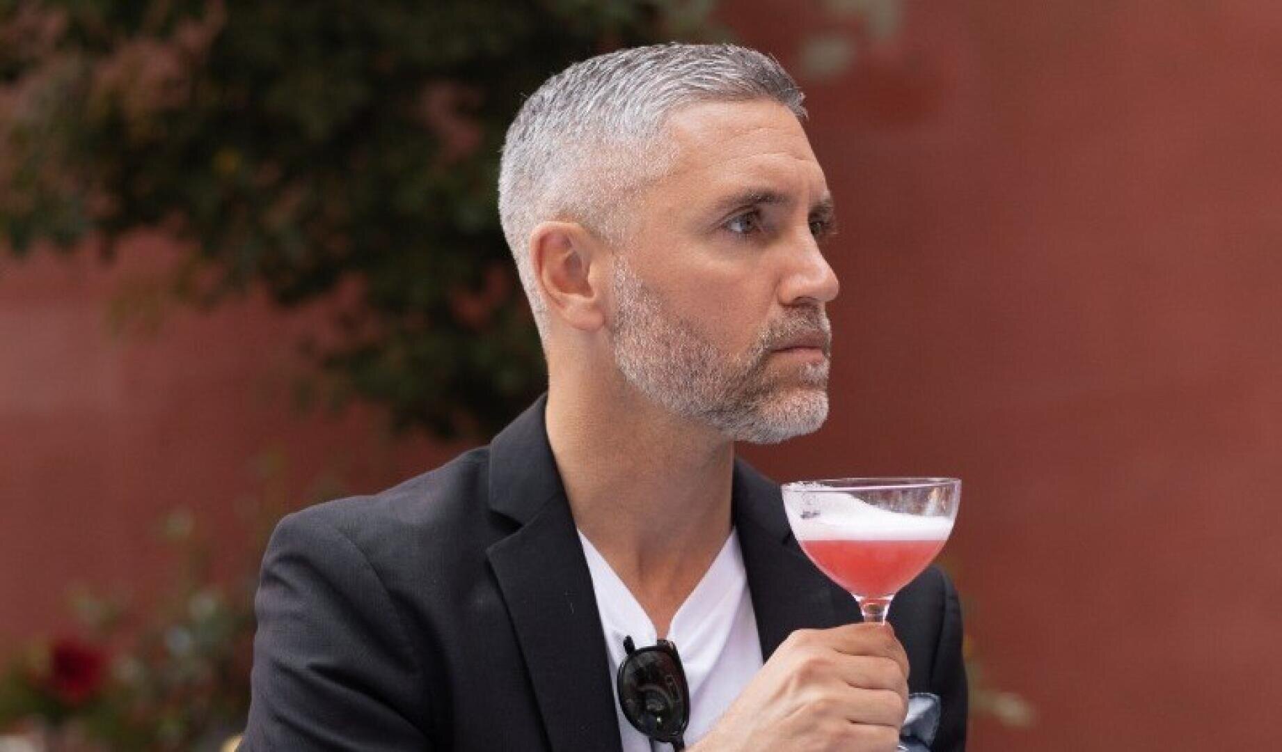 Ein Mann mit kurzen grauen Haaren und Bart, der einen schwarzen Blazer und ein weißes Hemd trägt, hält einen rosafarbenen Cocktail in einem Coupe-Glas und blickt nach links vor einem unscharfen Hintergrund im Freien.