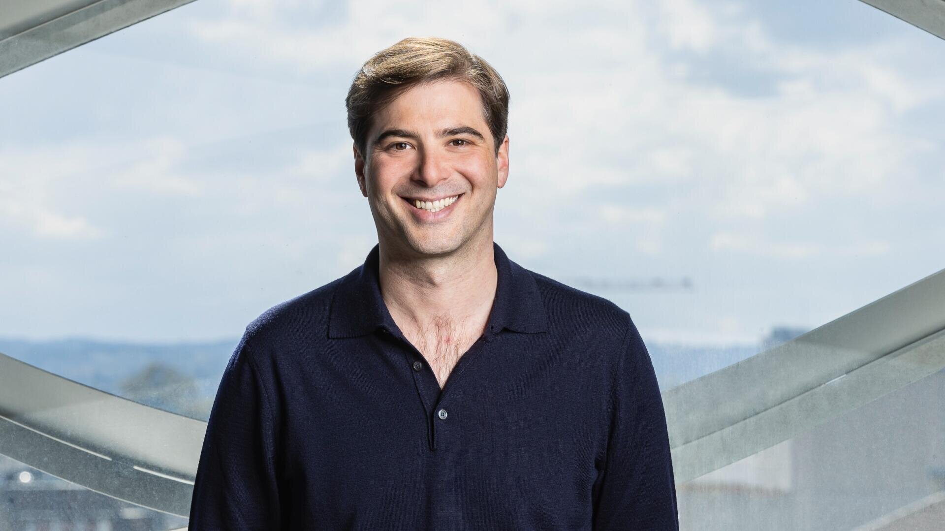 Ein Mann mit hellbraunem Haar und einem marineblauen Polohemd lächelt, während er in einem Innenraum vor großen Fenstern mit einem hellen, bewölkten Himmel im Hintergrund steht.