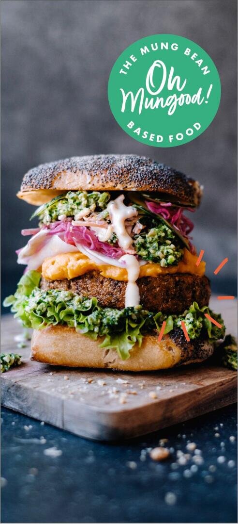 Ein dickes, gestapeltes veganes Sandwich mit Salat, eingelegten Zwiebeln, einem pflanzlichen Patty, Soße und einem Mohnbrötchen liegt auf einem Holzbrett. Auf einem grünen Etikett steht: The Mung Bean Oh Mungood! Essen auf pflanzlicher Basis.