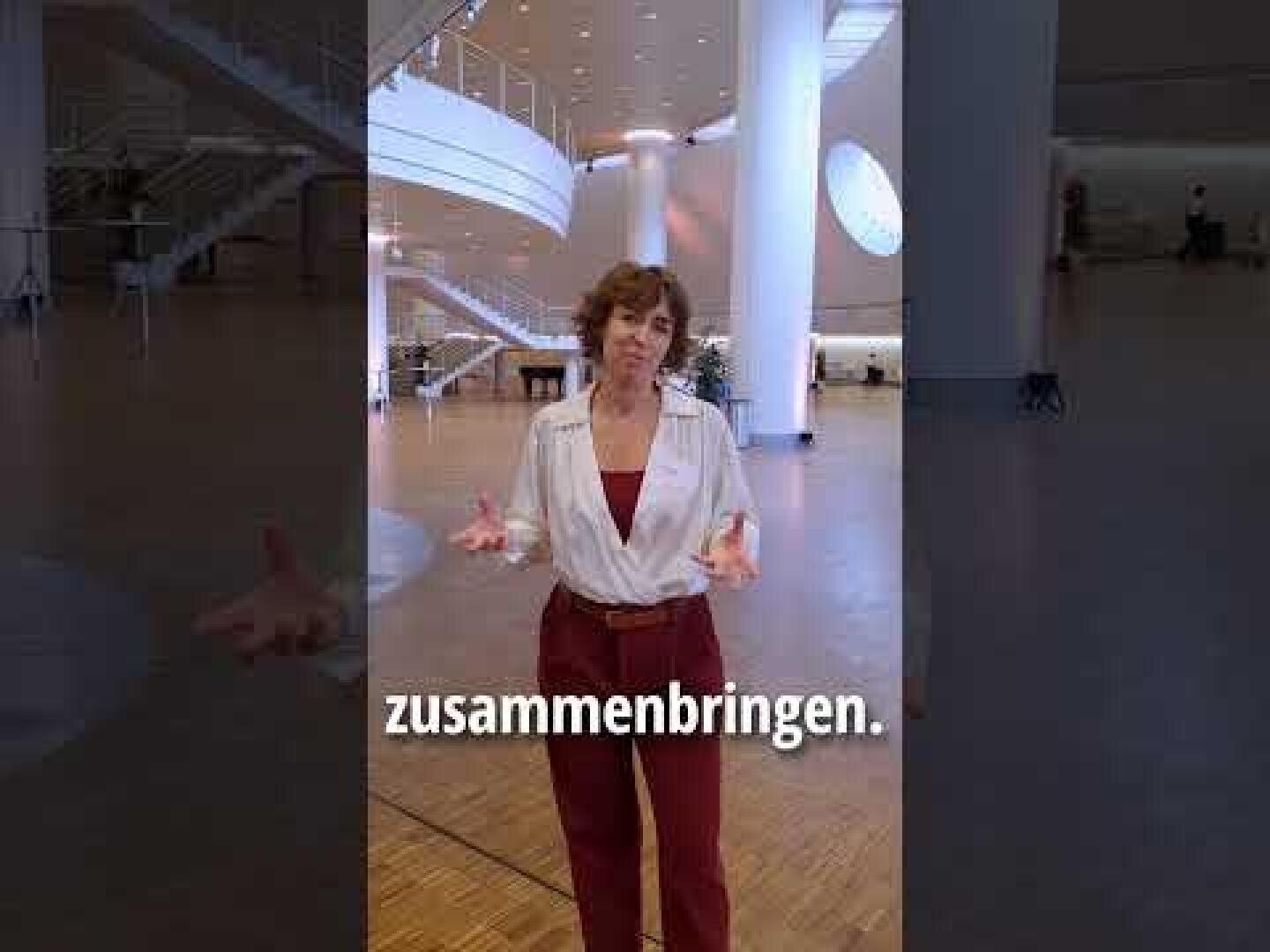 Eine Frau mit kurzen Haaren, die eine weiße Bluse und eine rote Hose trägt, steht in einem geräumigen, modernen Gebäude mit hohen Säulen und Holzfußboden. Das deutsche Wort "zusammenbringen" erscheint quer über dem Bild.