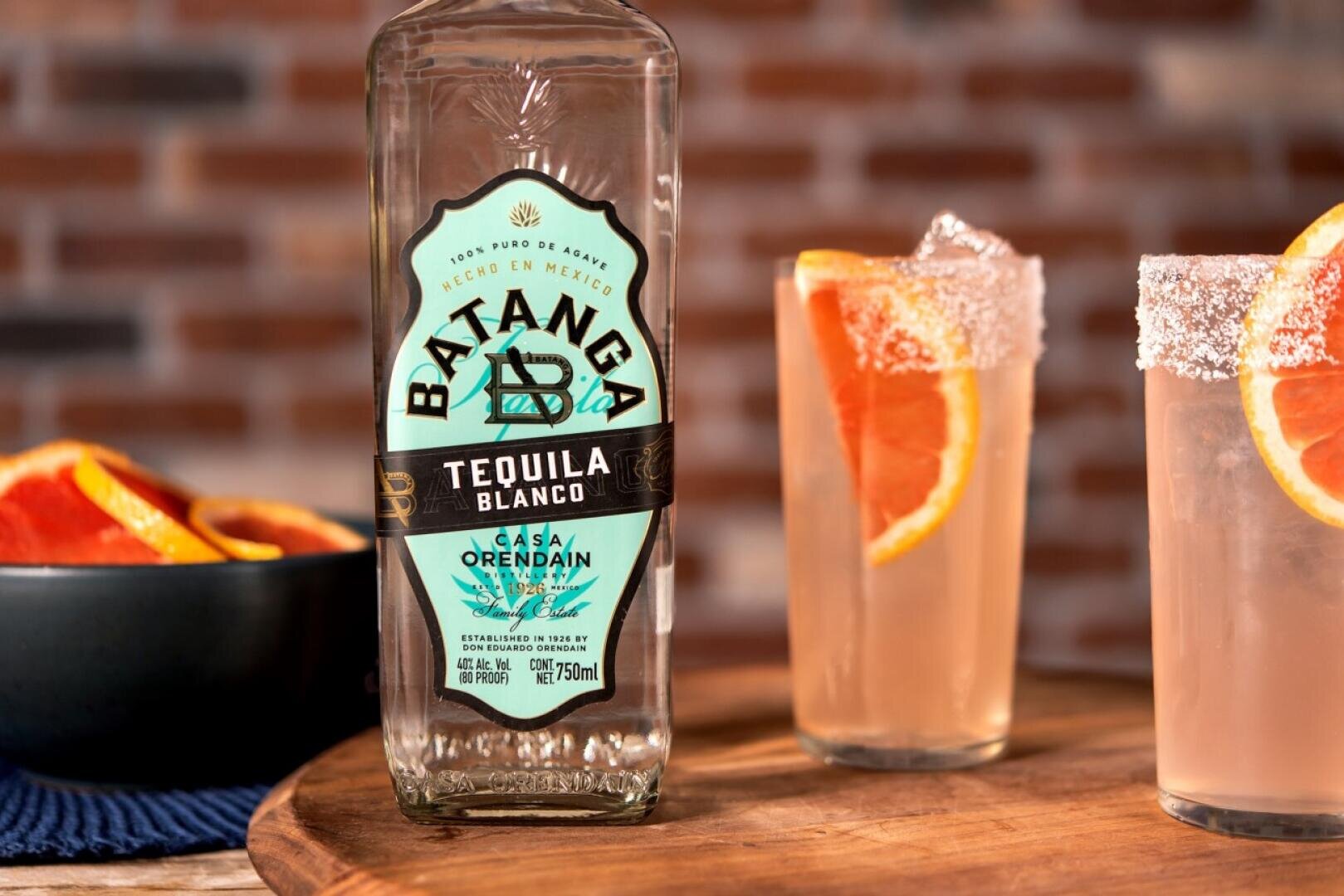 Eine Flasche Batanga Tequila Blanco steht auf einer Holzplatte neben zwei Cocktails, die mit Orangenscheiben und Salzrändern garniert sind. Im Hintergrund befindet sich eine Schale mit Orangenscheiben, hinter denen eine Backsteinmauer verschwimmt.