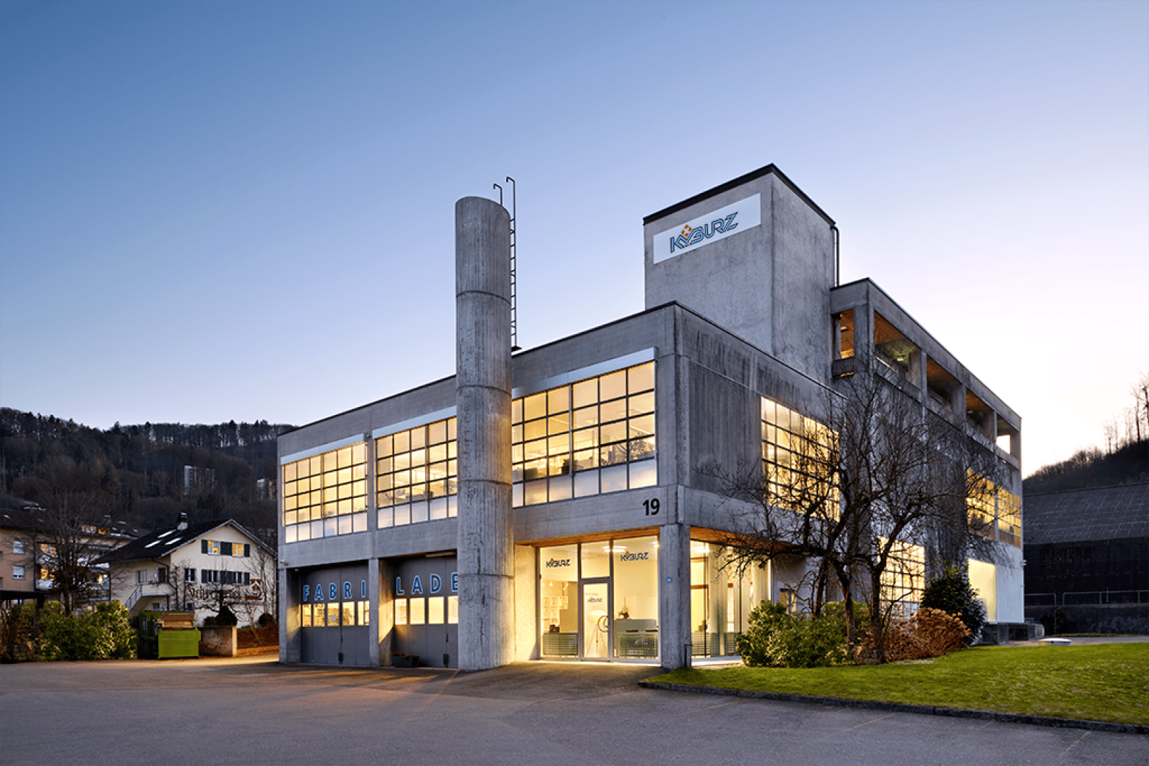 Un bâtiment rectangulaire moderne en béton, doté de grandes fenêtres et baigné de lumière, se dresse au bord d'une route au crépuscule. L'enseigne KREATIVEZ et le numéro 19 sont inscrits à l'extérieur. Des arbustes entourent l'entrée.