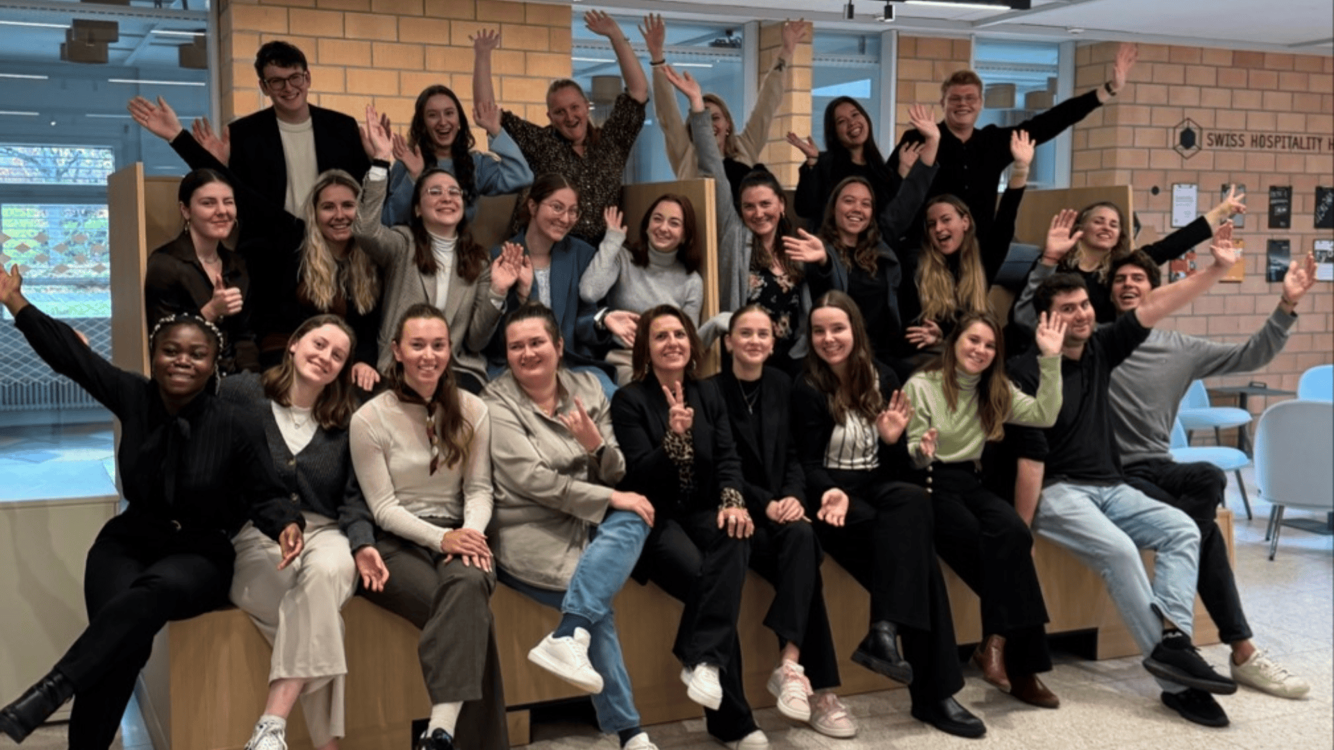 Un groupe d'environ 25 jeunes adultes souriants posent dans un espace intérieur, assis et debout sur des sièges étagés, et agitent leurs mains vers l'appareil photo avec enthousiasme. Ils semblent détendus et heureux dans un espace moderne et lumineux.
