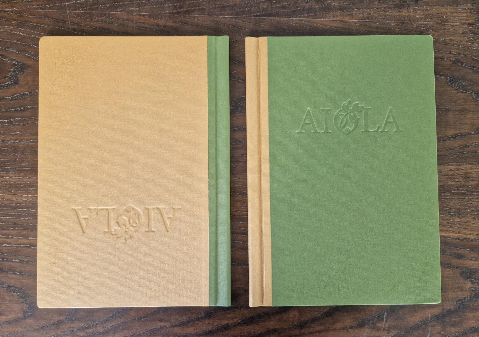 Zwei Hardcover-Bücher mit AIOLA-Prägung auf dem Einband liegen auf einer Holzfläche. Das linke Buch ist hellbraun und steht auf dem Kopf, während das rechte Buch grün ist und auf dem Kopf steht.