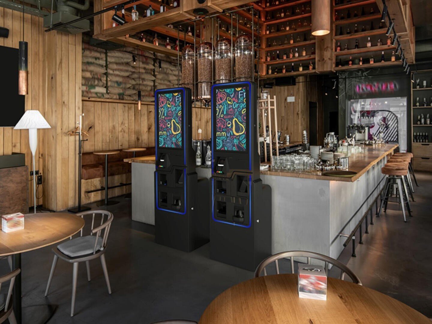 Ein modernes Restaurant mit Holzwänden und -möbeln, das zwei digitale Selbstbedienungskioske mit bunten Bildschirmen in der Nähe des Barbereichs bietet. Der Raum ist gut beleuchtet und stilvoll dekoriert.