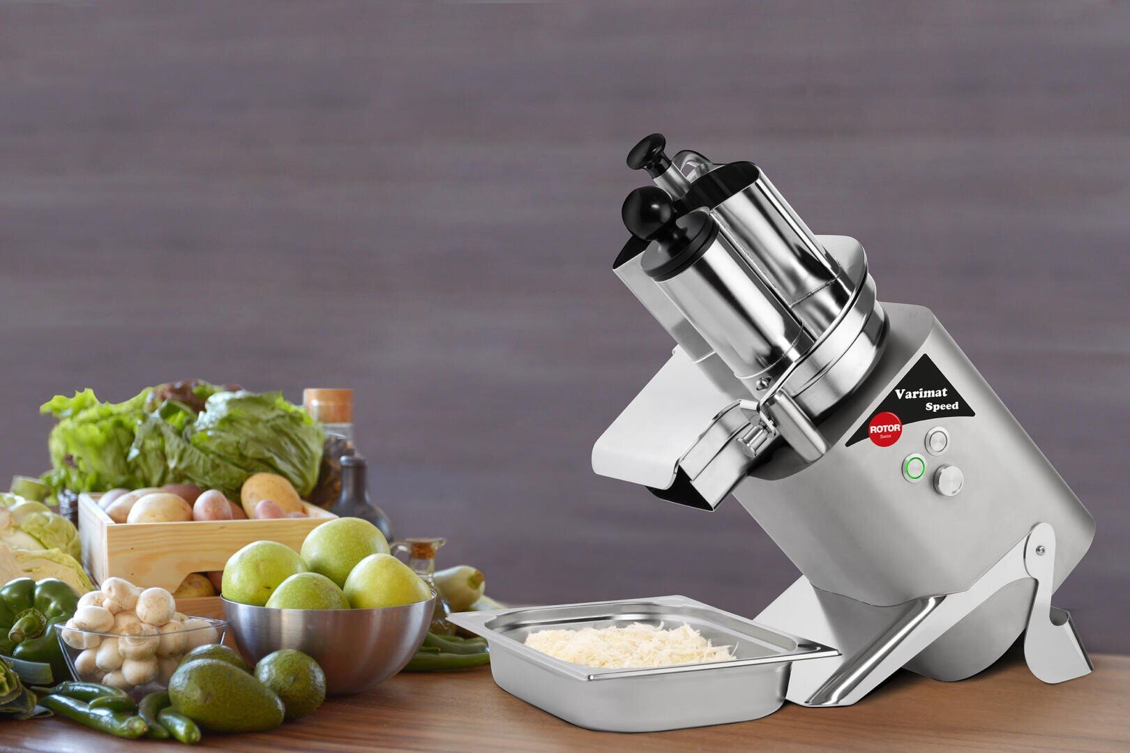 Un robot ménager commercial est posé sur un comptoir à côté d'un plateau de fromage râpé et de légumes frais, notamment de la laitue, des pommes, des champignons, des poivrons verts et des tomates.