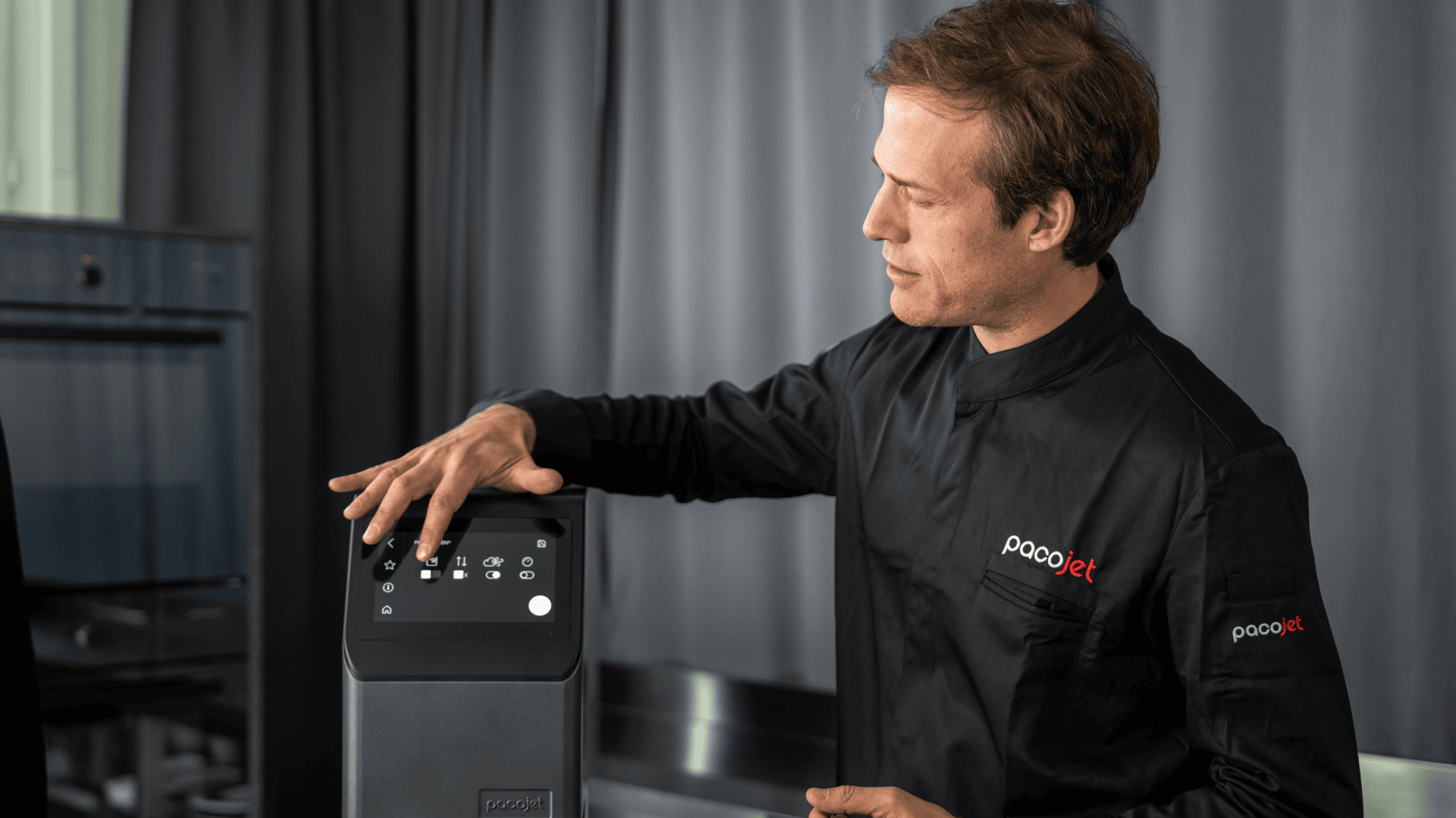 Un cuisinier en uniforme Pacojet noir fait fonctionner une machine Pacojet et appuie sur des touches du panneau de commande, tandis qu'un rideau sombre et des ustensiles de cuisine sont visibles à l'arrière-plan.