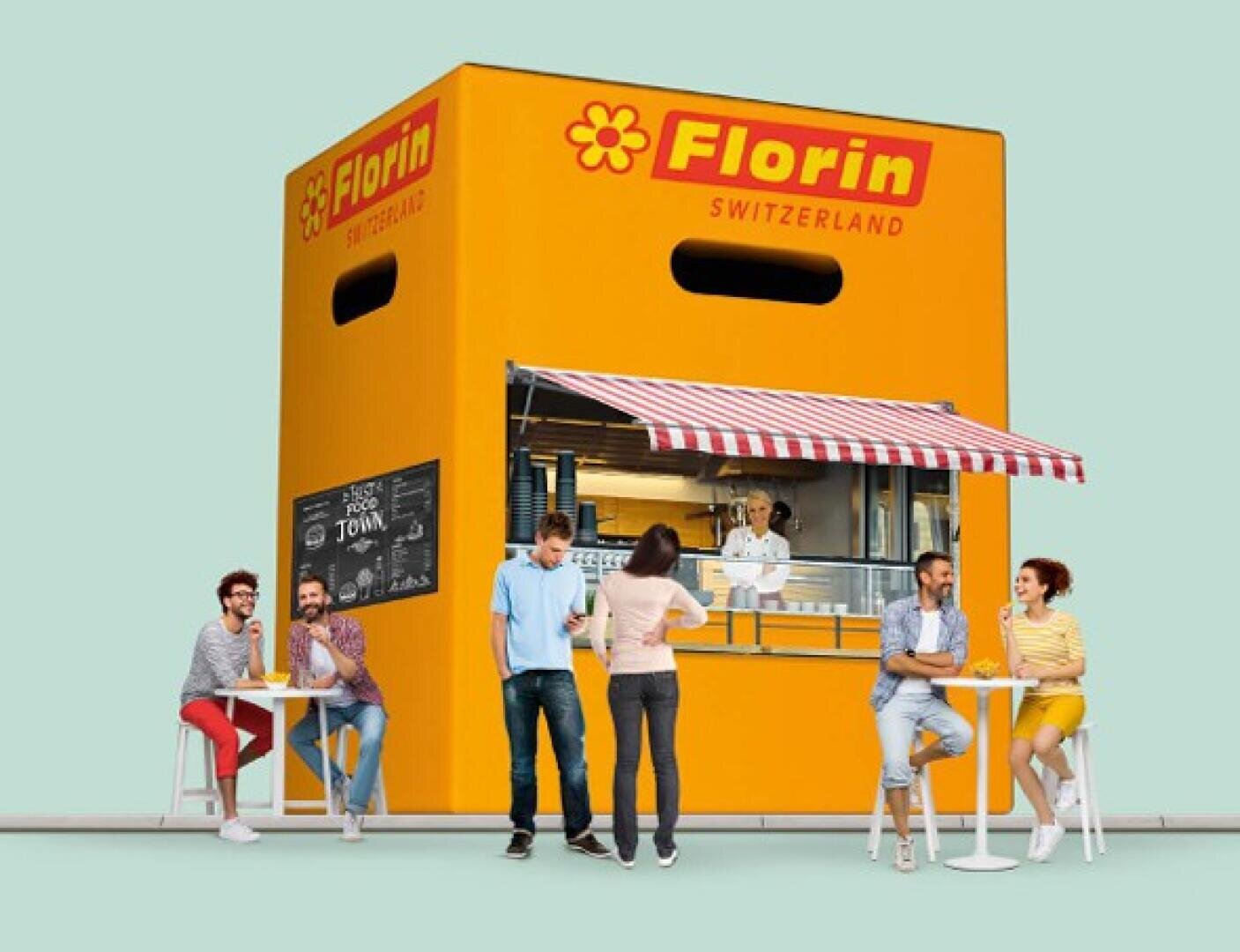 Un groupe de personnes déguste des snacks et des boissons devant un stand de nourriture en forme d'énorme emballage de fromage jaune Florin Switzerland. A l'intérieur du stand, on peut voir un cuisinier sous un auvent à rayures rouges et blanches.