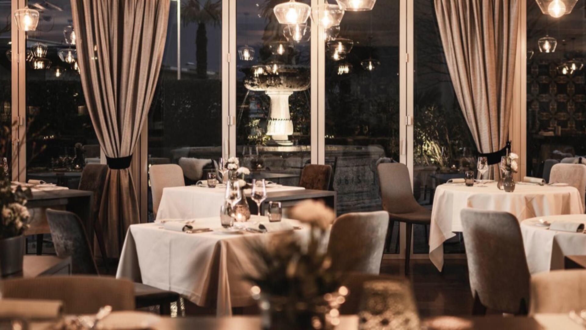 Eleganter Speisesaal eines Restaurants mit gedeckten Tischen, weißen Tischtüchern und gedämpfter Beleuchtung. Große Fenster mit Vorhängen geben den Blick auf einen beleuchteten Springbrunnen und Grünflächen frei und schaffen ein warmes, einladendes Ambiente.