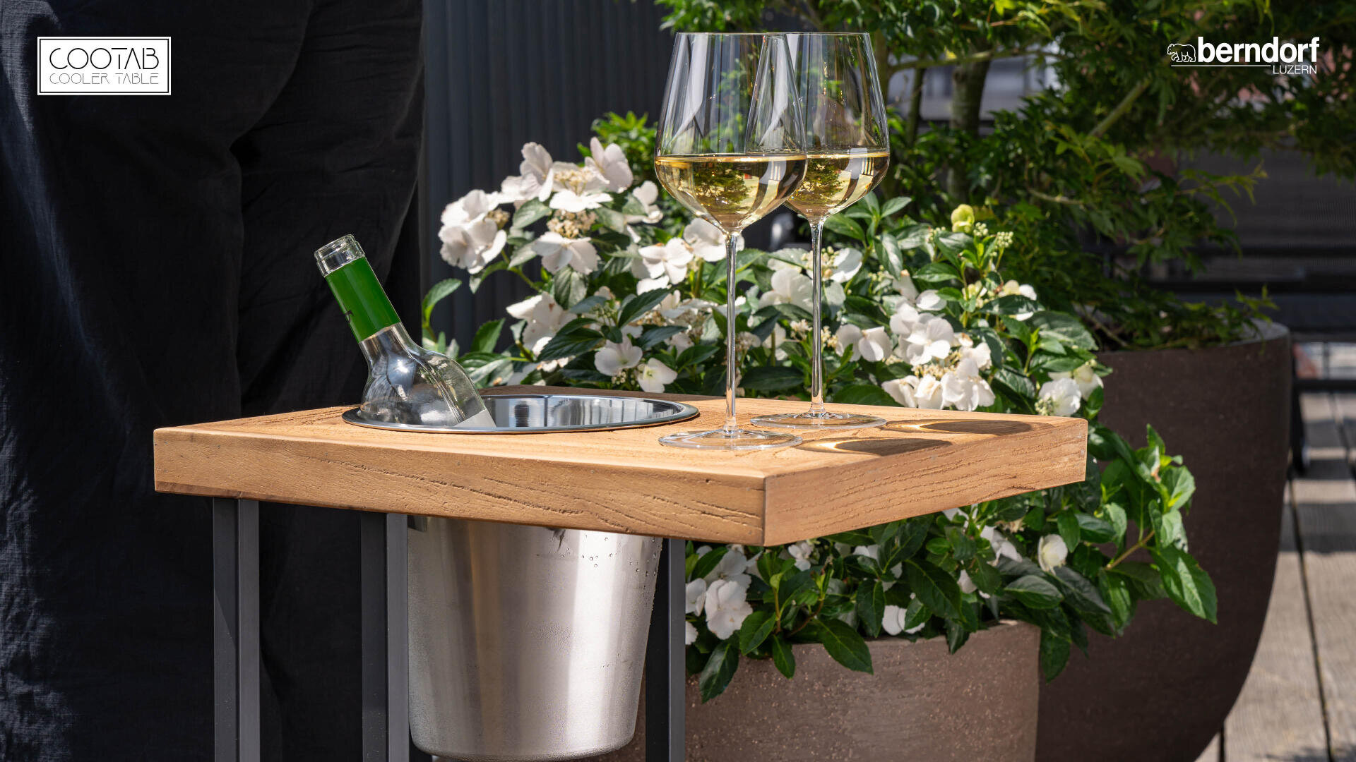 Une table en bois avec un seau à glace en métal intégré contenant une bouteille de vin et deux verres de vin blanc, entourée de plantes vertes et de fleurs blanches en plein air.