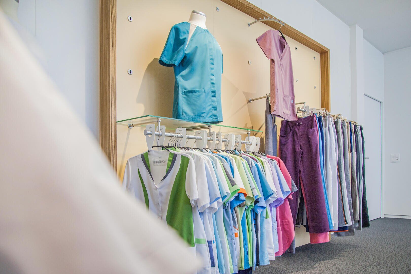In einem gut beleuchteten Raum sind verschiedene bunte medizinische Kittel und Uniformen auf Bügeln und Schaufensterpuppen an einer Wand ausgestellt, die eine Reihe von Stilen und Farben zeigen.